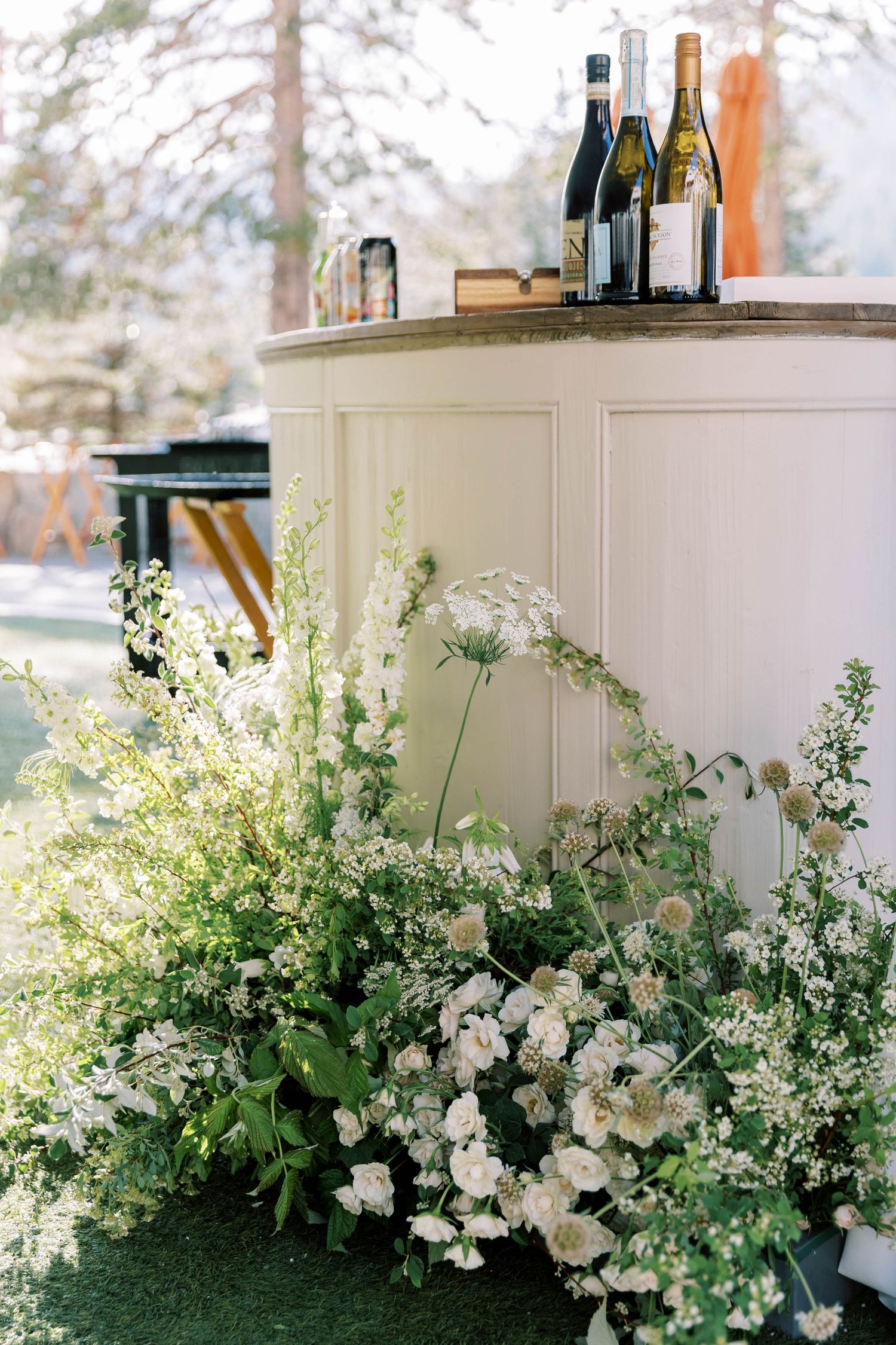 White wedding bar setup with wine bottles and romantic floral arrangements on artificial grass.