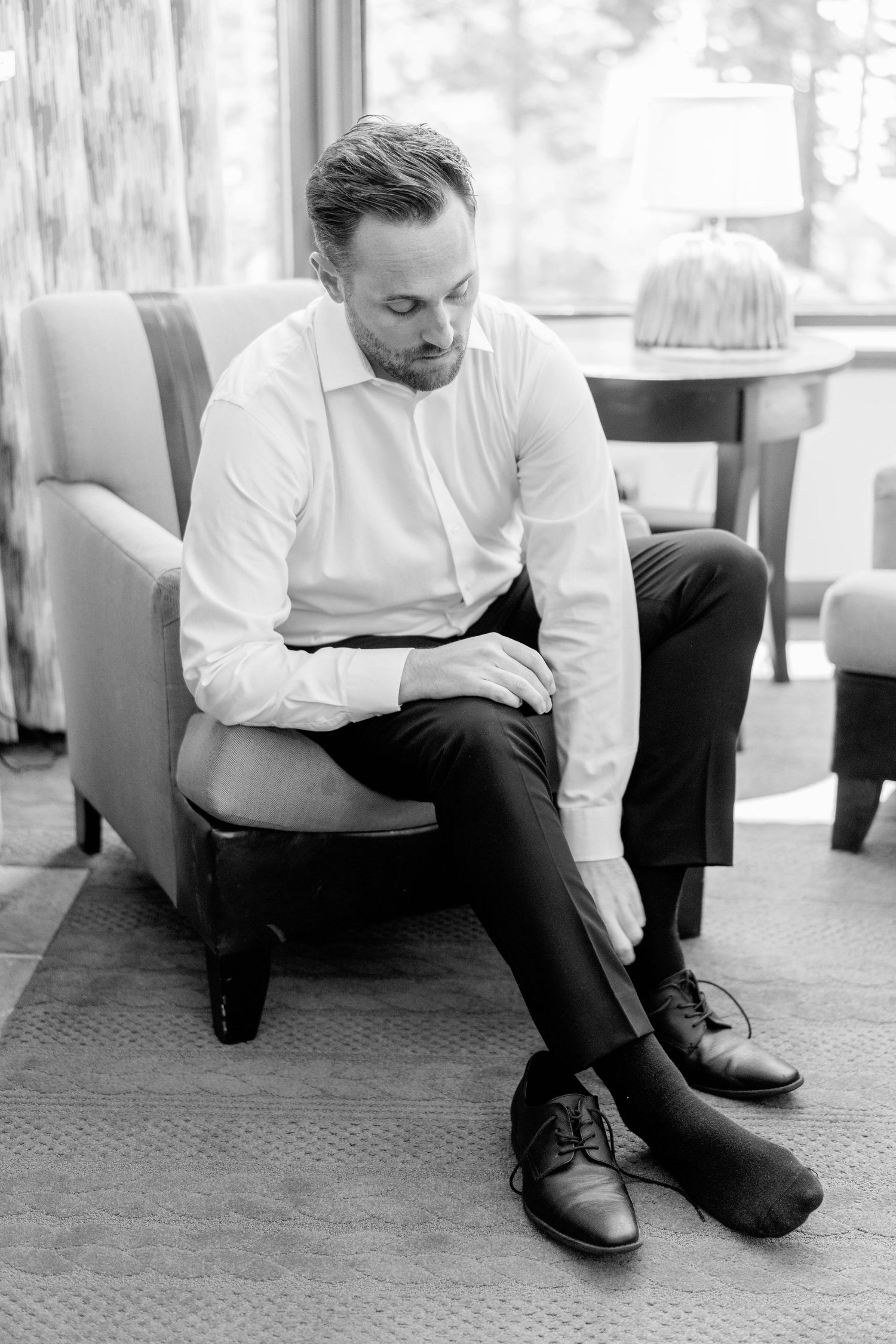 Groom in formal attire sits thoughtfully in an upholstered chair while getting ready.