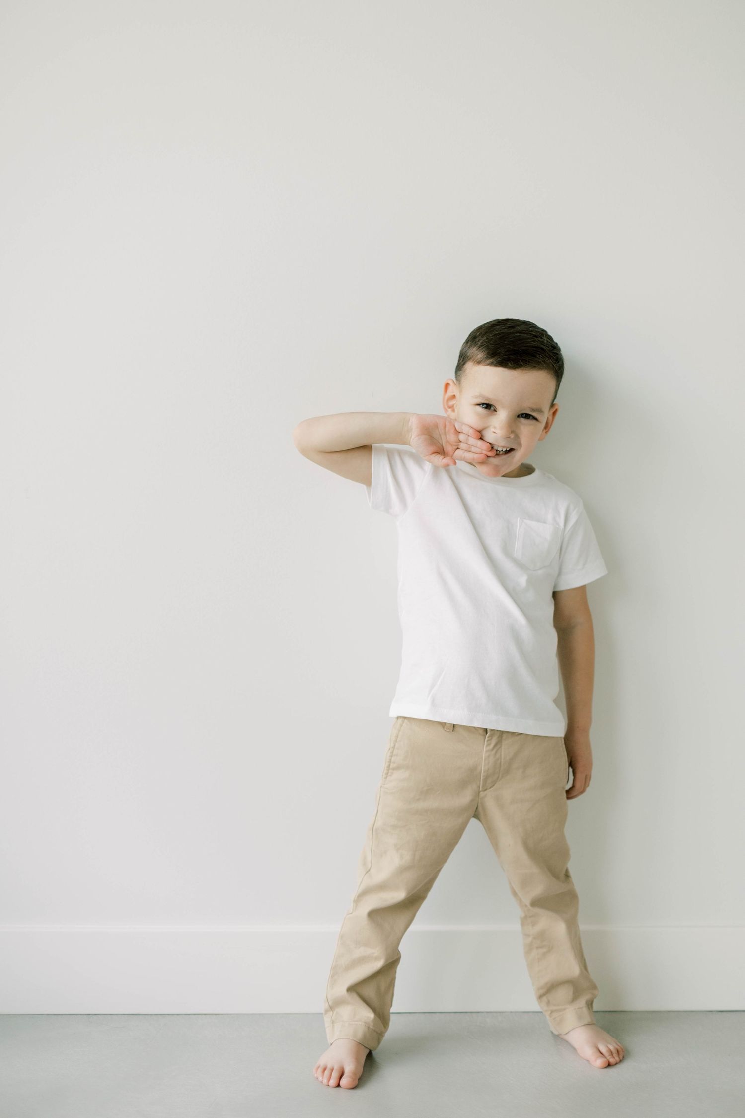 Child in white t-shirt and khaki pants poses playfully against white wall.
