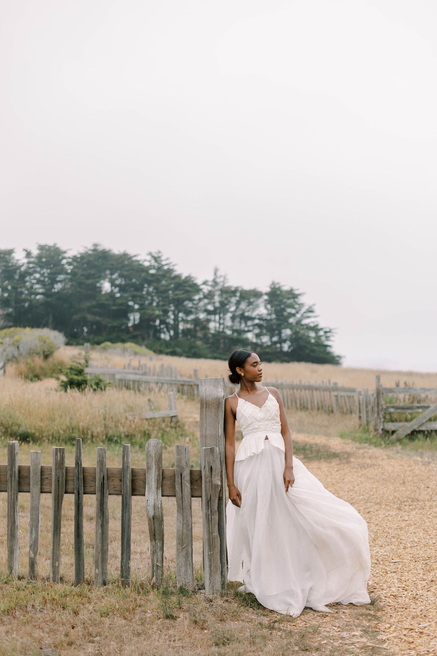 A flowing white wedding dress moves gracefully against a rustic wooden fence along the coastline.