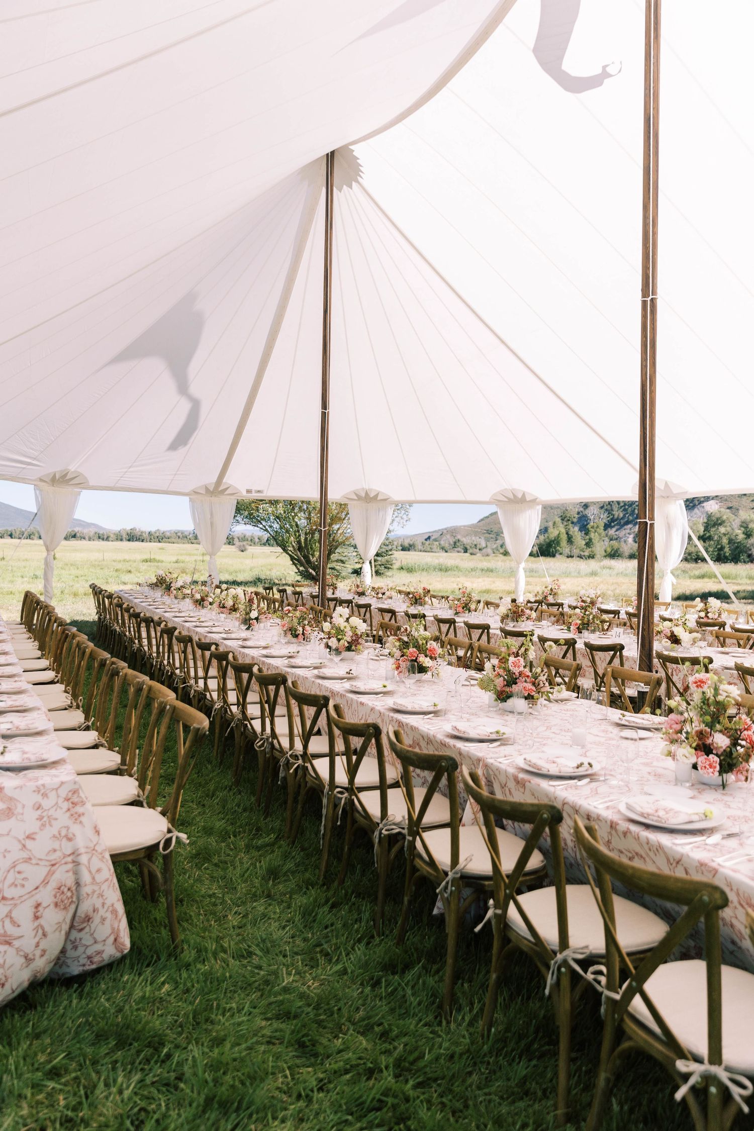 Elegant outdoor wedding reception setup with long banquet tables under white tent canopy and rustic wooden cross-back chairs.