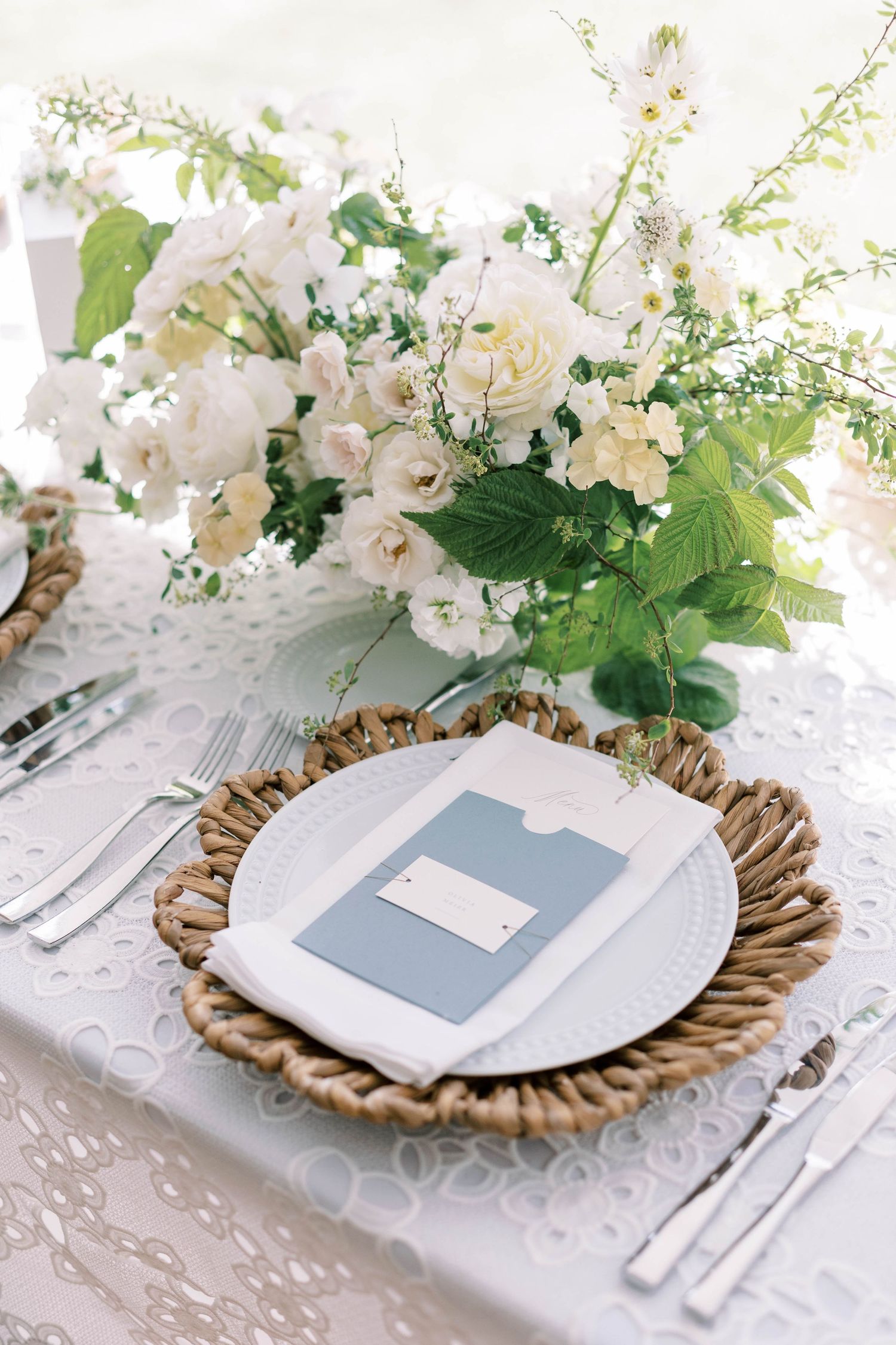Place setting with white floral centerpiece and blue menu card on gold-rimmed charger plate.