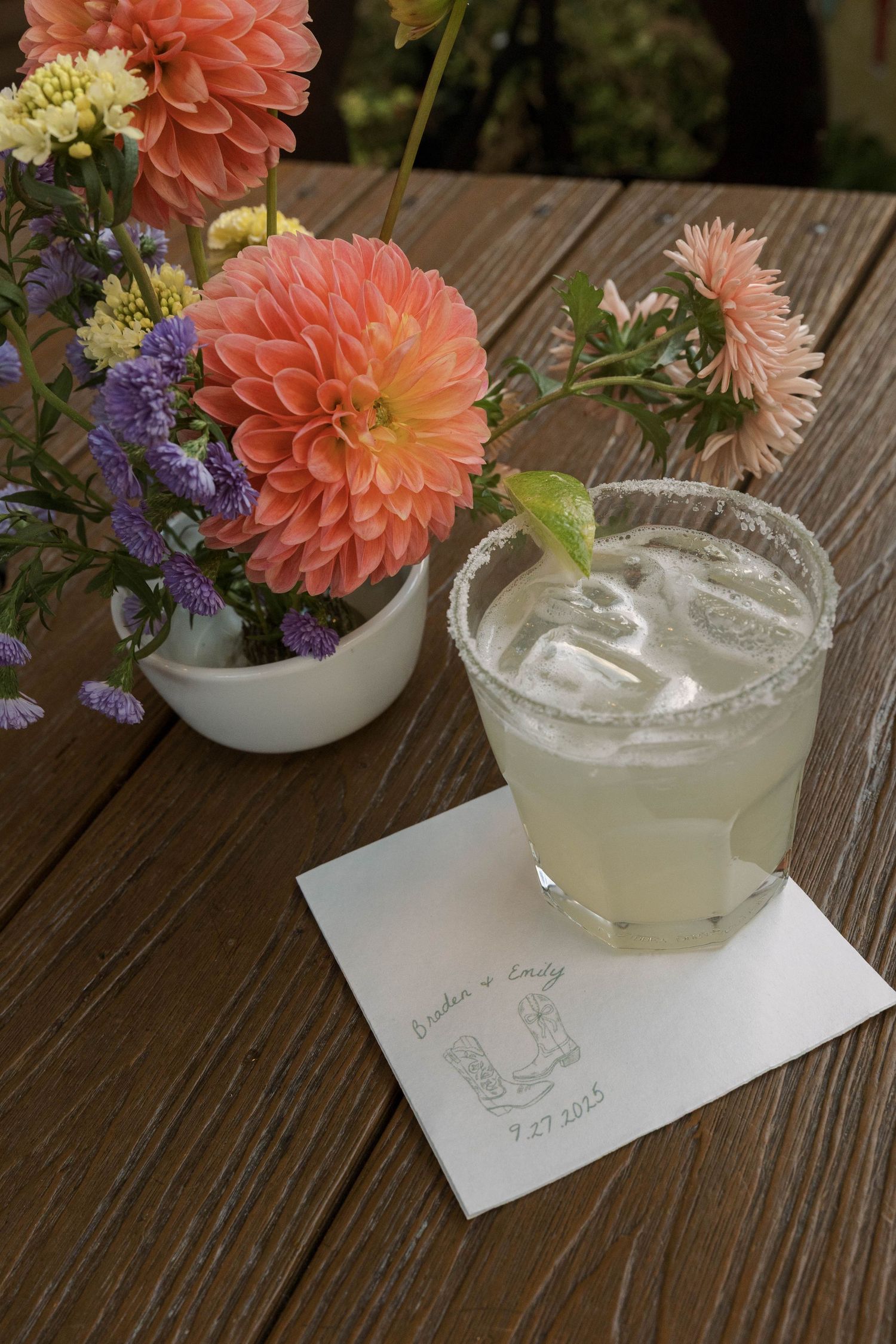 Elegant cocktails garnished with coral dahlias and purple flowers sit on a rustic wooden table with handwritten notes.