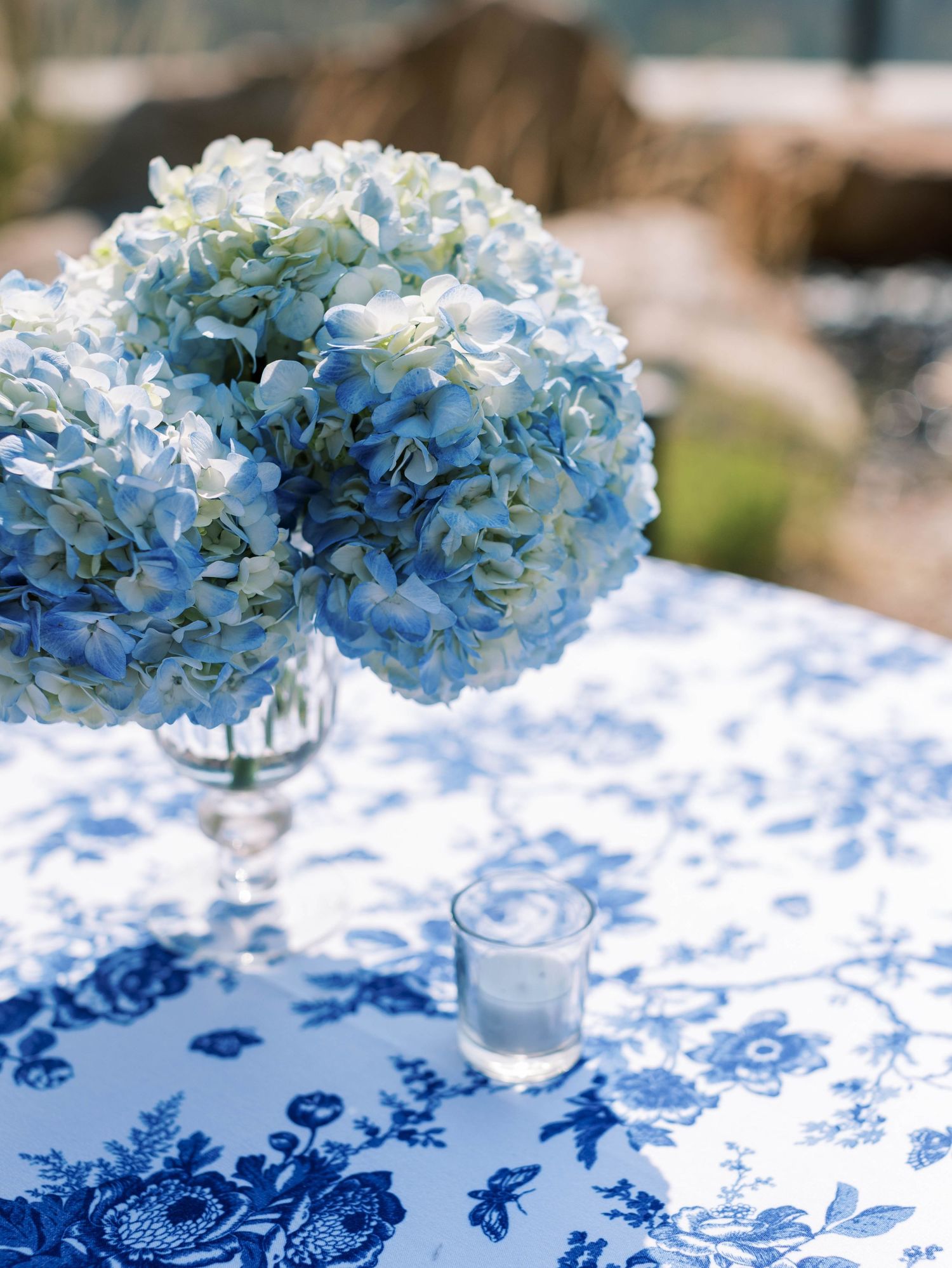Blue hydrangea flower arrangements in glass vases on a white and blue floral patterned tablecloth create an elegant display.