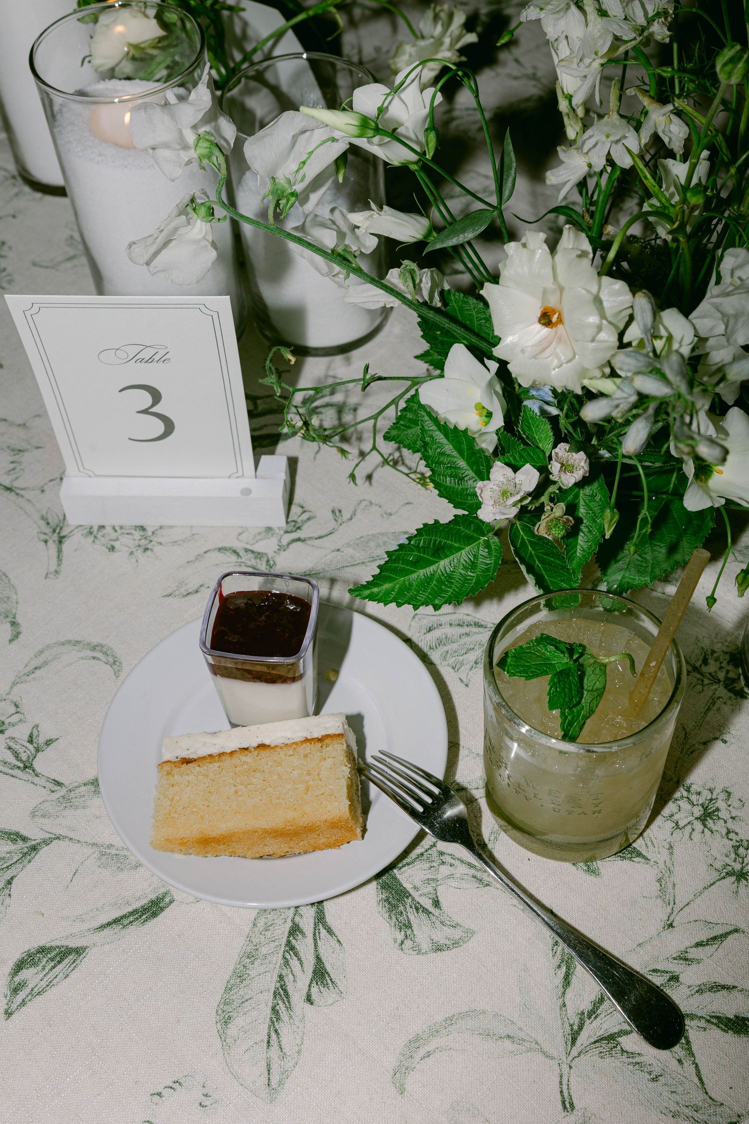 Elegant wedding table setting with white floral centerpieces, table numbers, and dessert plates on floral linens.