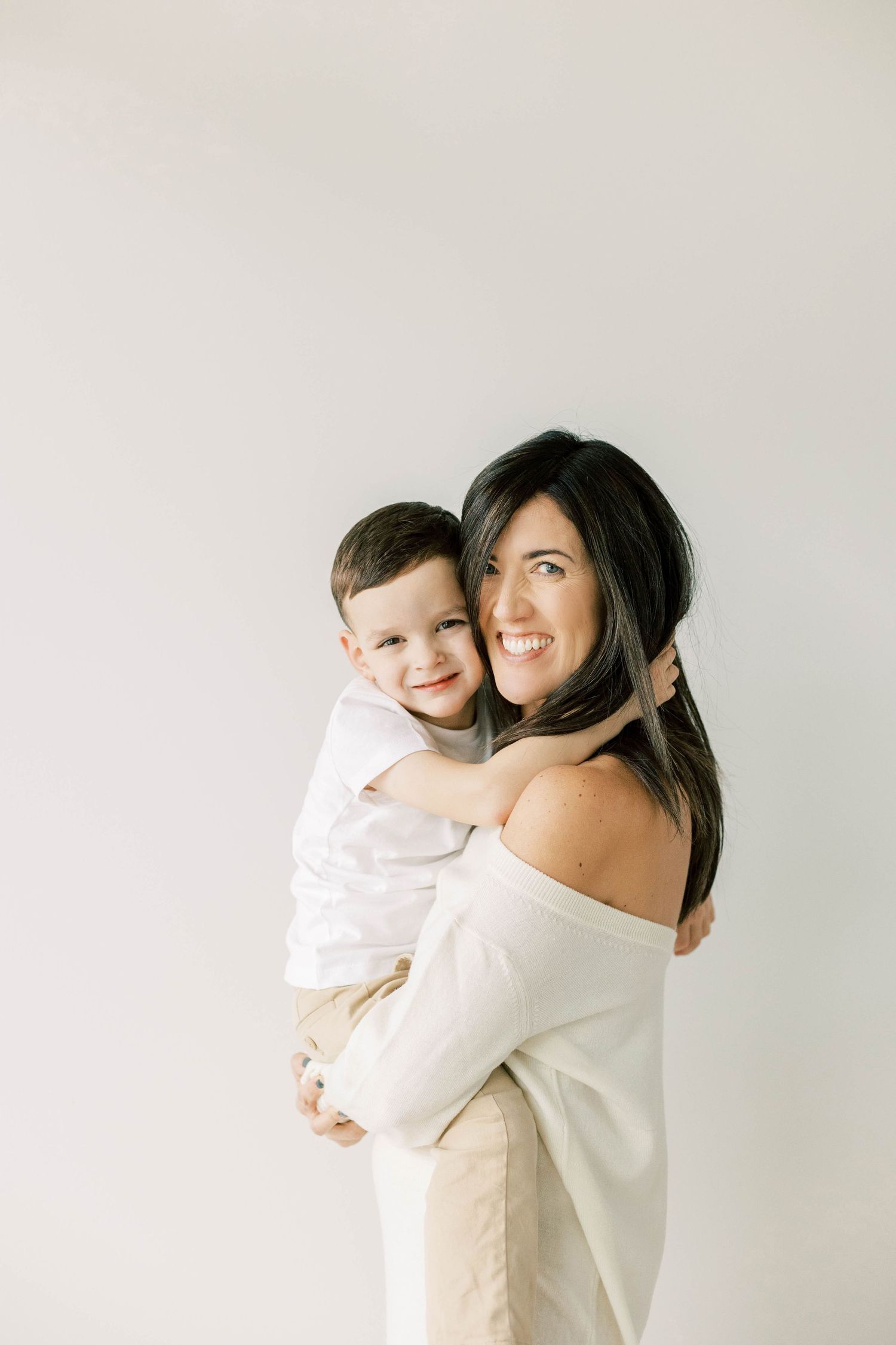 Young mother in white sweater holding smiling toddler against neutral background.