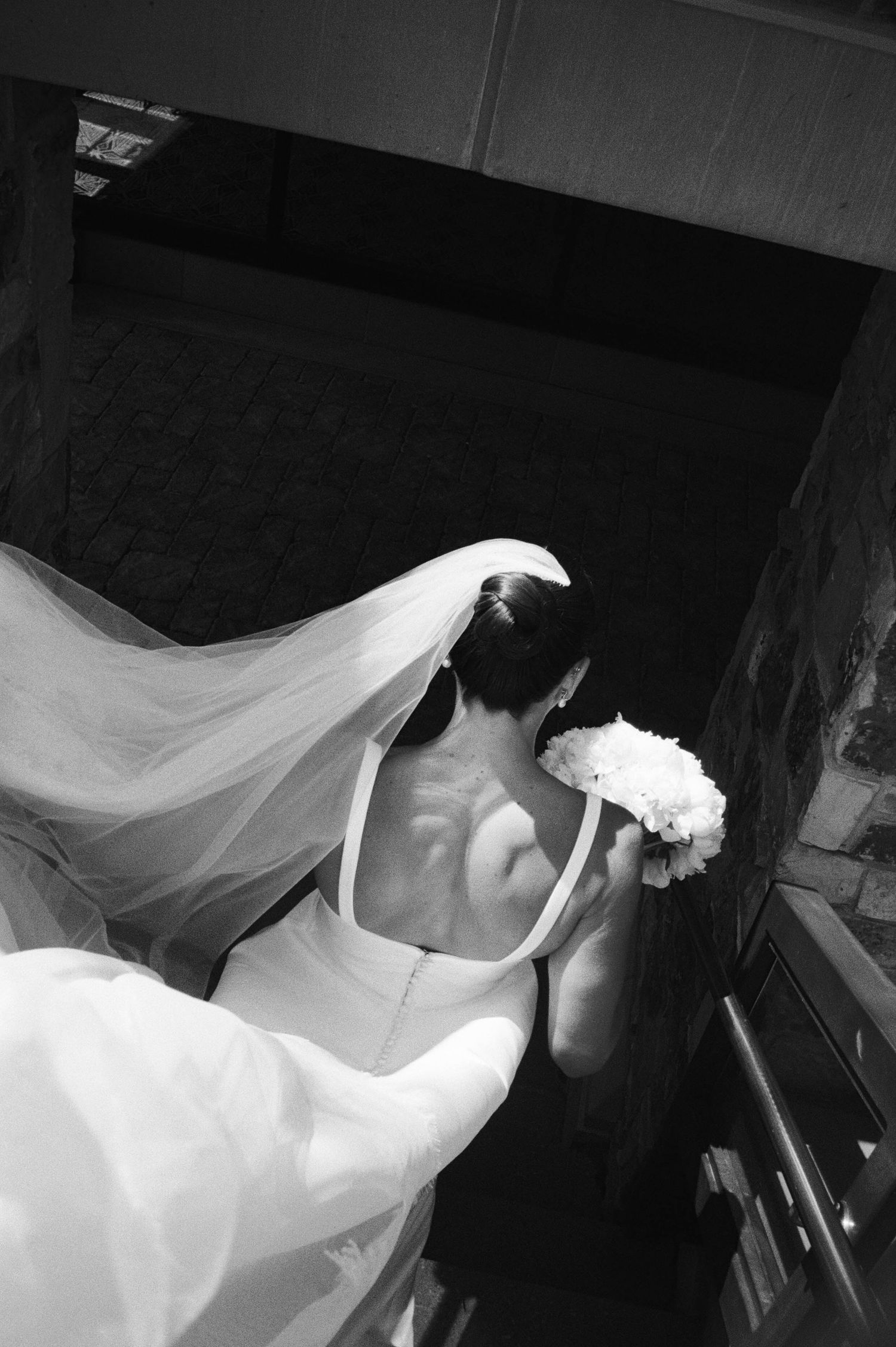 A bride in a flowing white wedding dress and veil leans gracefully in a dramatic black and white photograph.