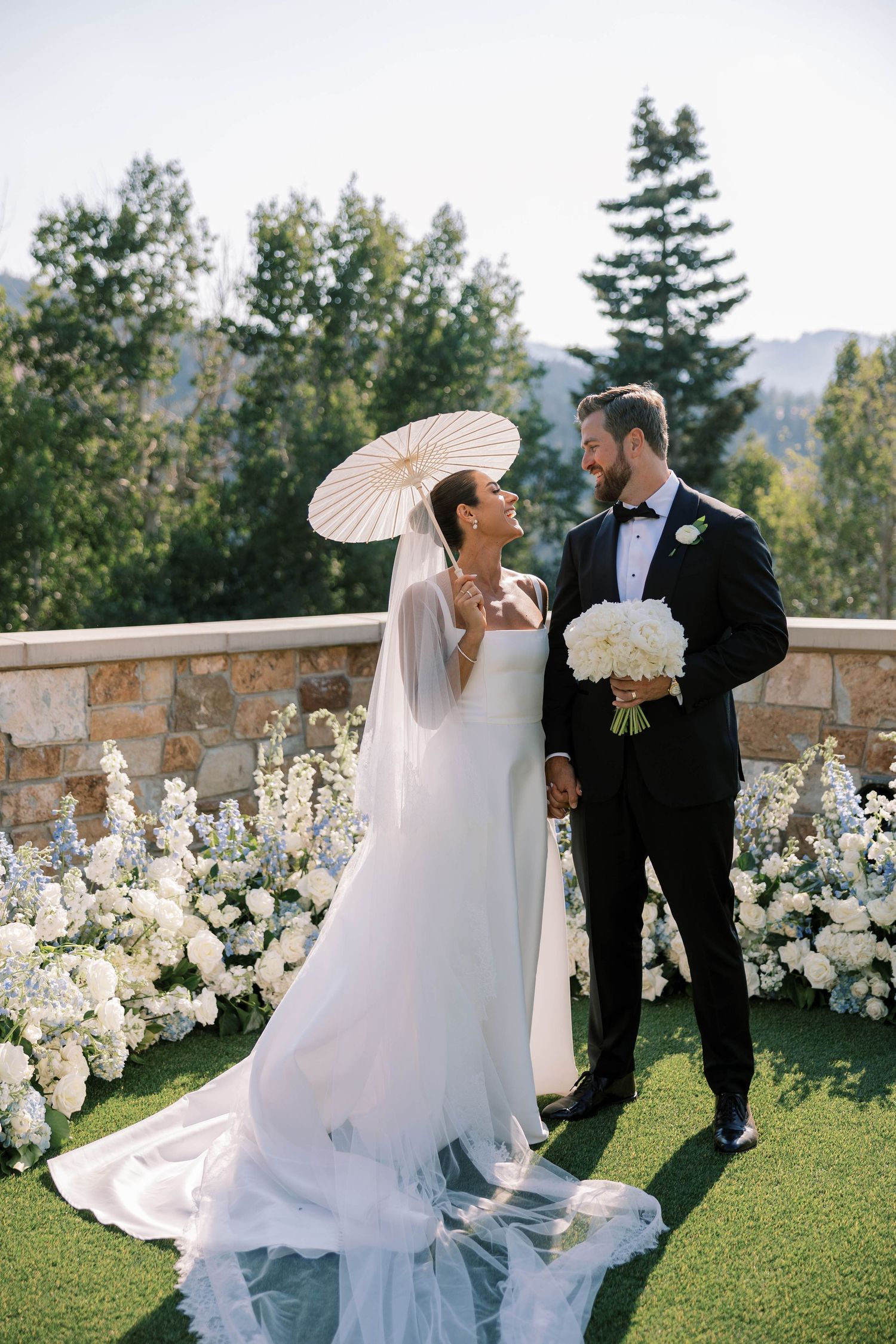 A romantic wedding couple poses on a grassy lawn with white floral decorations and mountain views in the background.