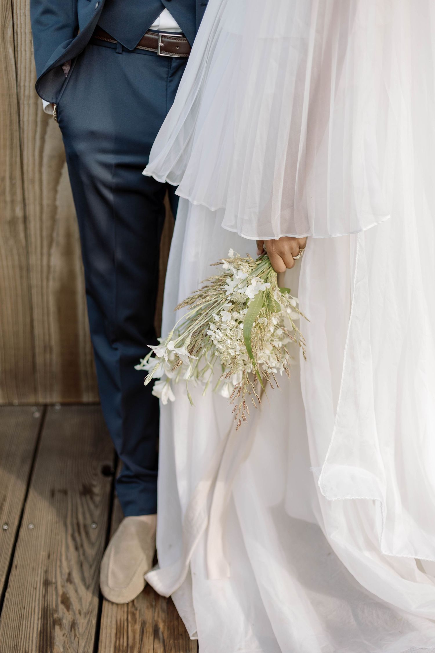 Close-up view of white wedding dress hem and navy suit pants with delicate white floral bouquet against wooden floor.