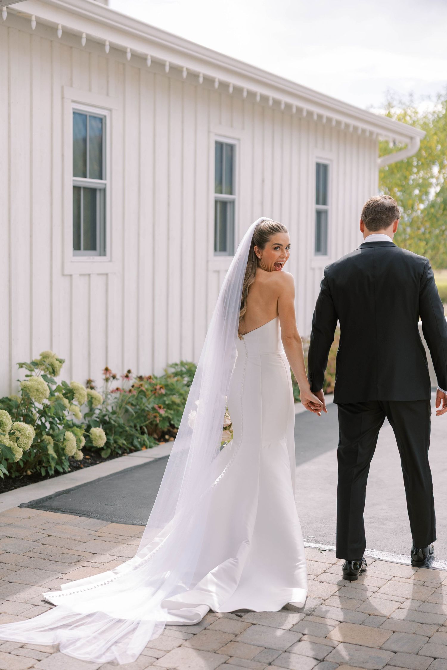 Elegant wedding couple stands by white barn building surrounded by hydrangea bushes.