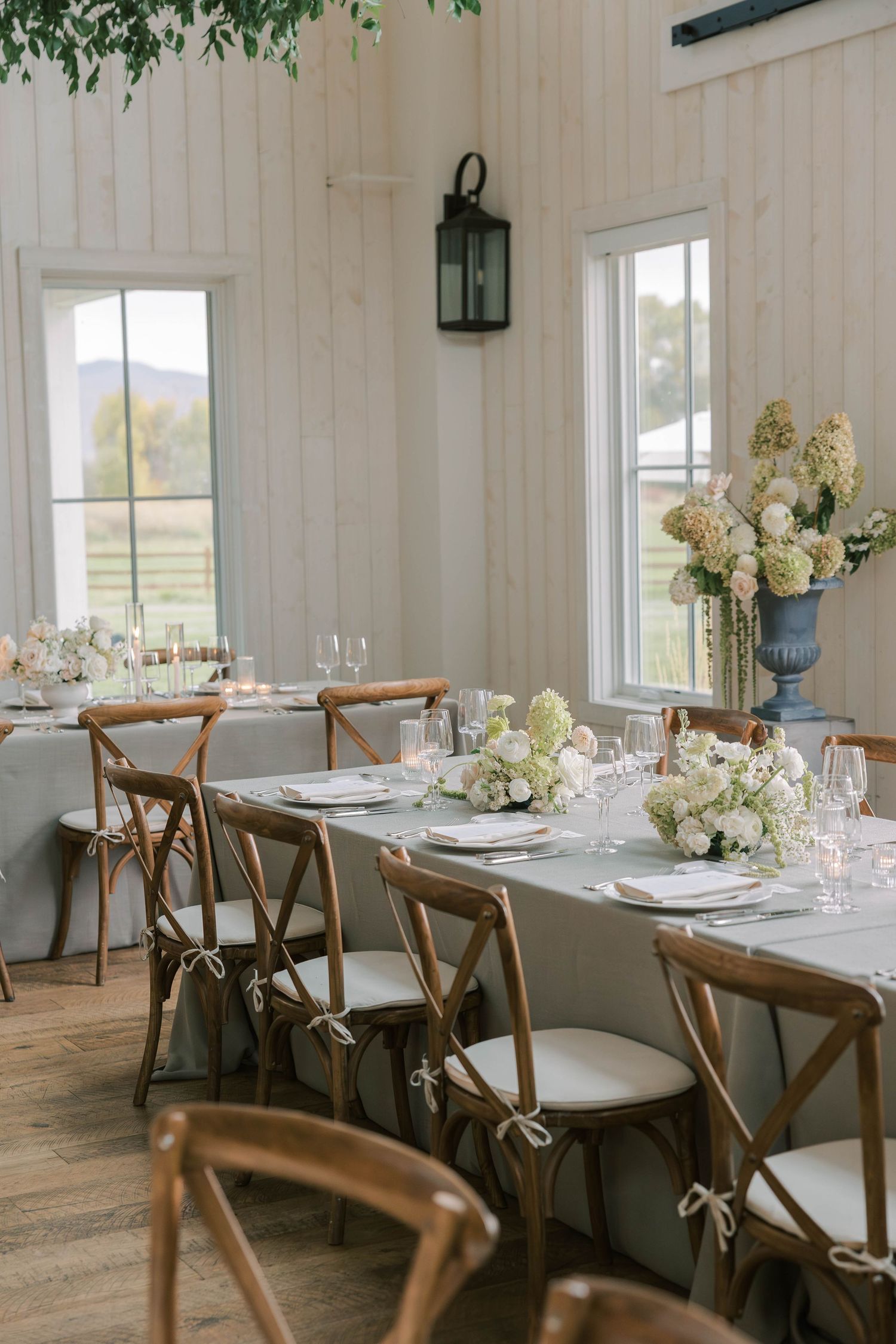 Elegant dining room with white walls, wooden crossback chairs, and white floral centerpieces set for a special occasion.