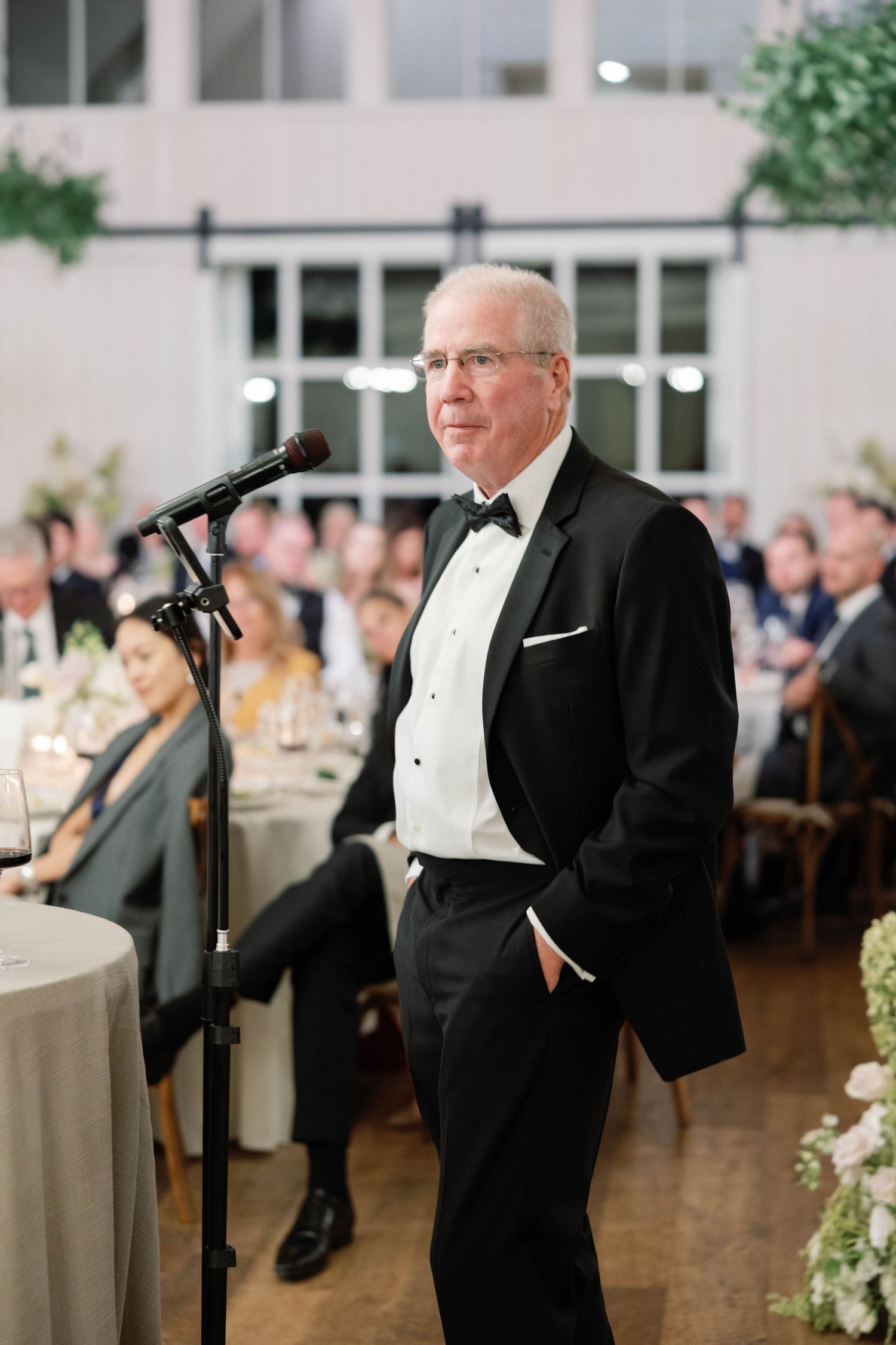 A speaker in a black tuxedo delivers a speech at an elegant indoor reception venue with guests seated in the background.