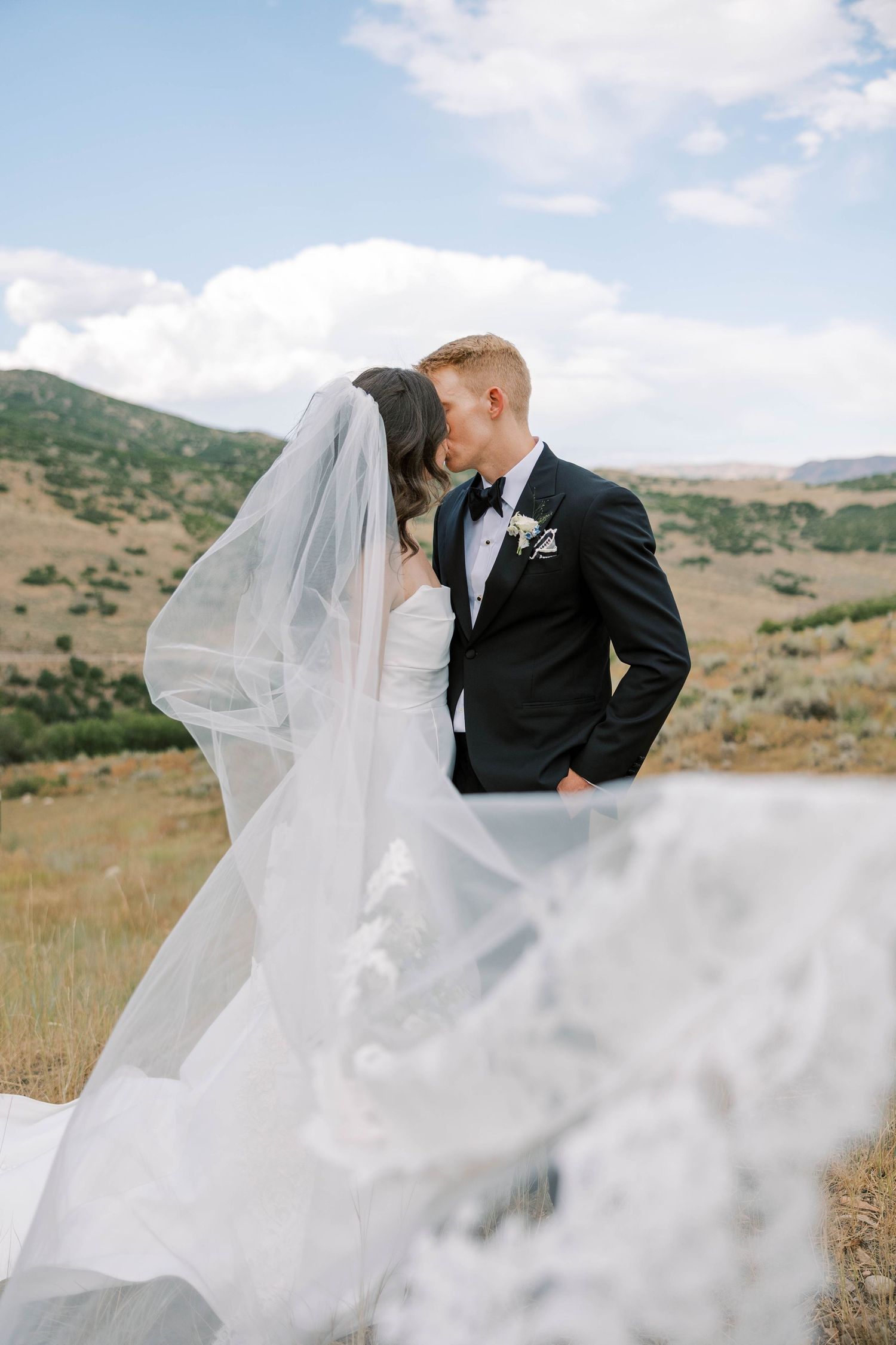 Elegant wedding couple photographed against scenic mountain landscape with flowing white veil.