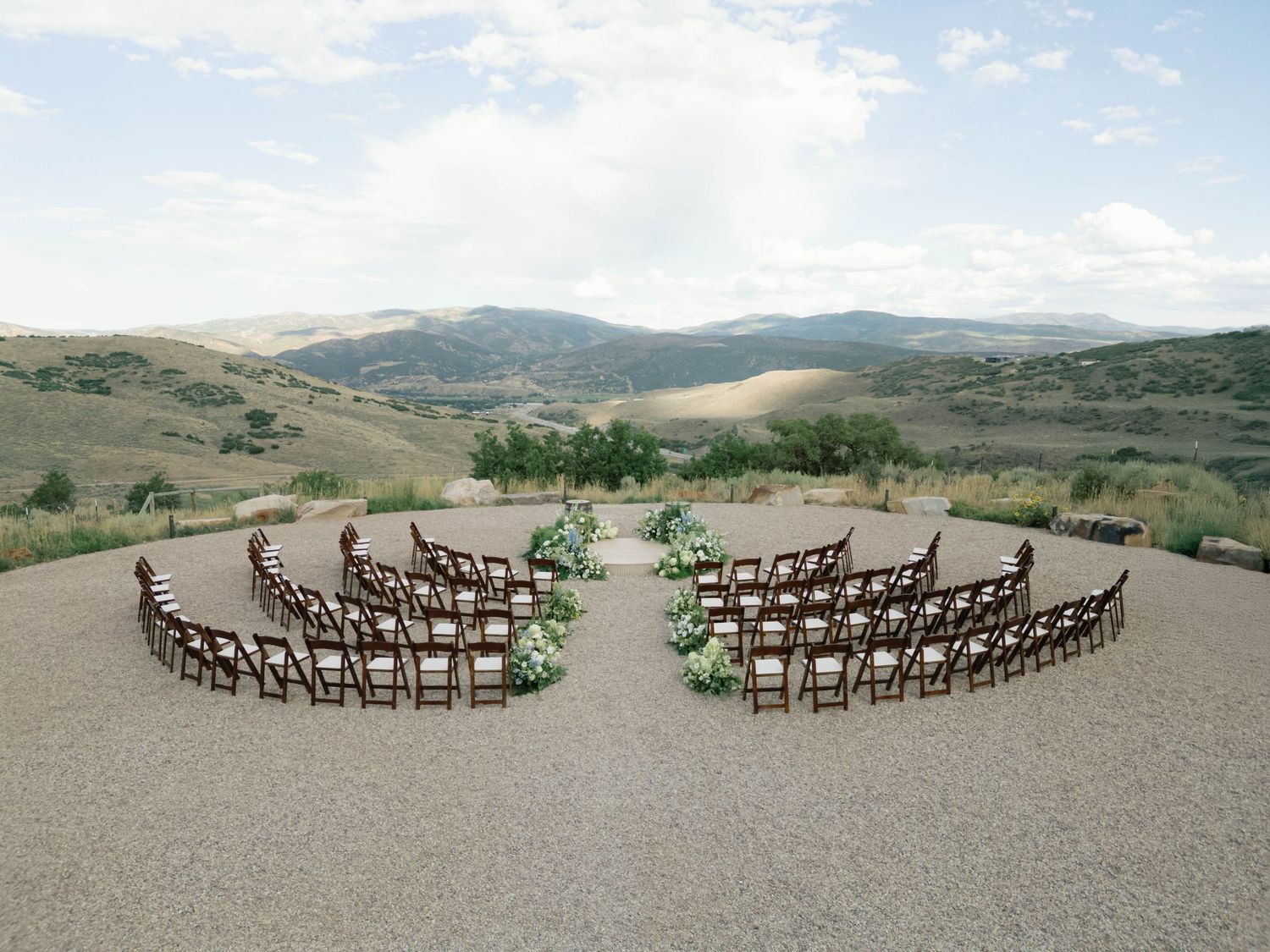Wooden chairs arranged in a curved semicircle formation on a hilltop overlooking rolling mountains in the distance.