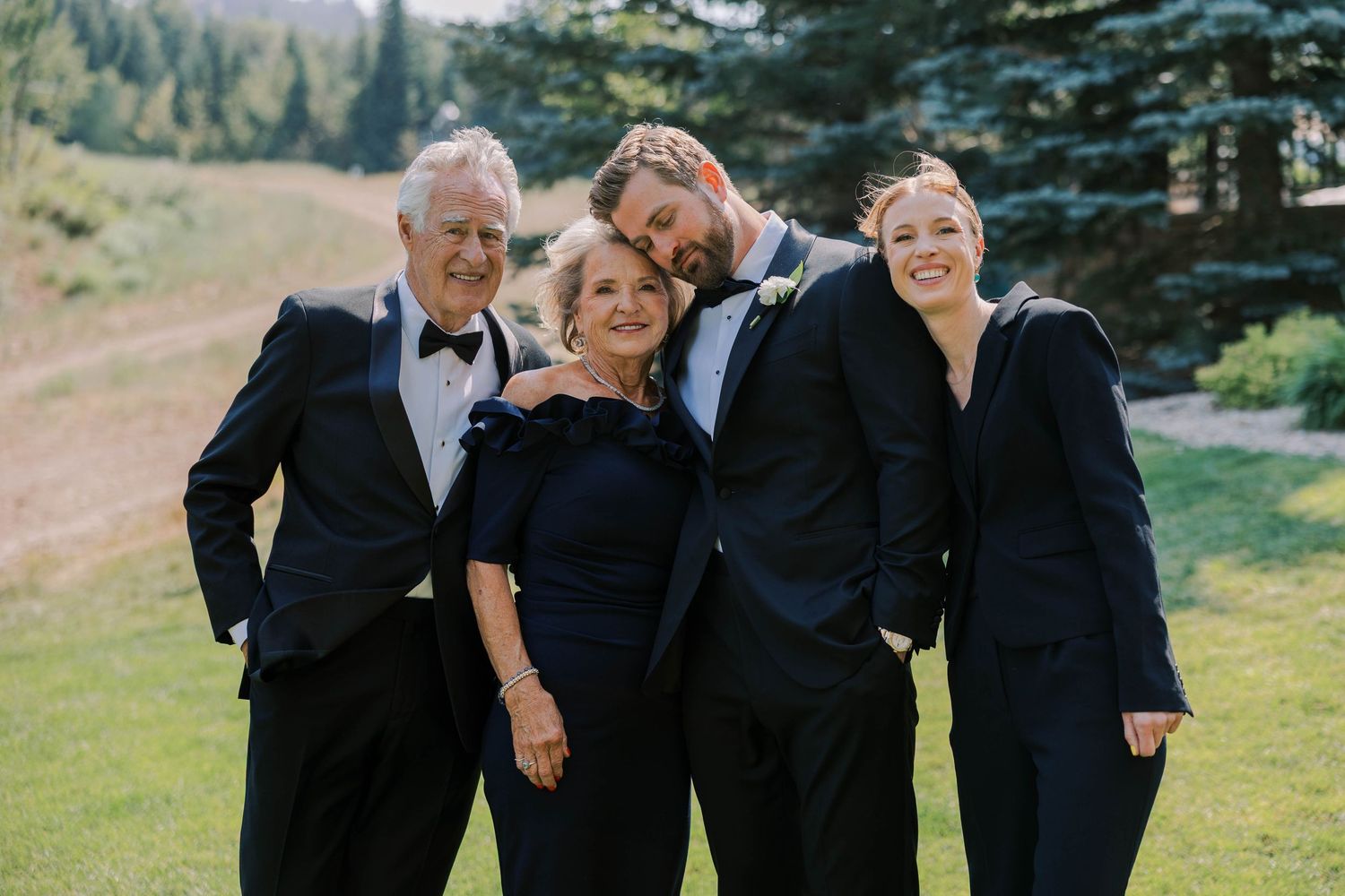 Wedding party in black attire laughing together outdoors with evergreen trees in background.