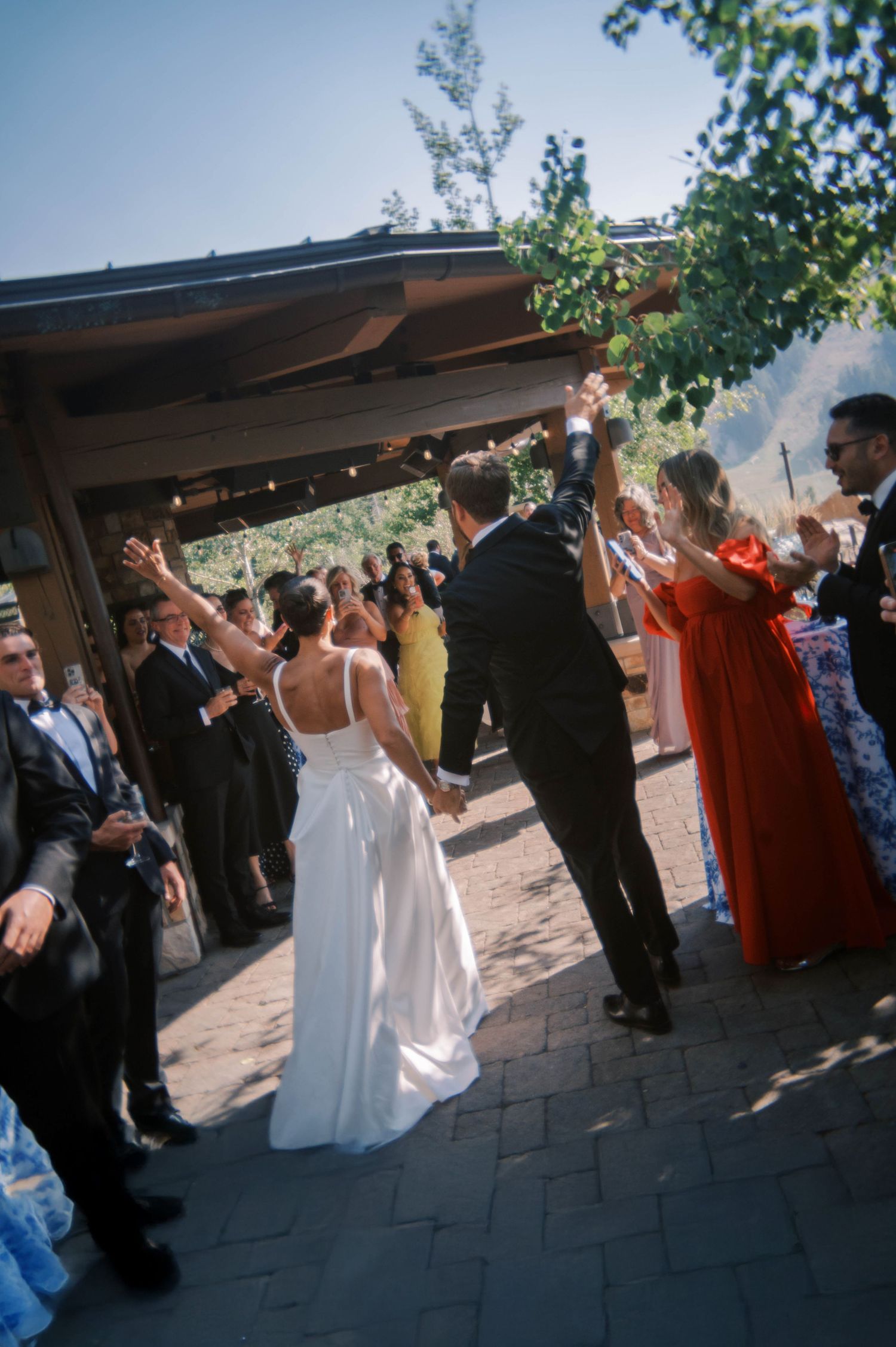 Wedding guests dance and celebrate at an outdoor reception under a covered patio area.
