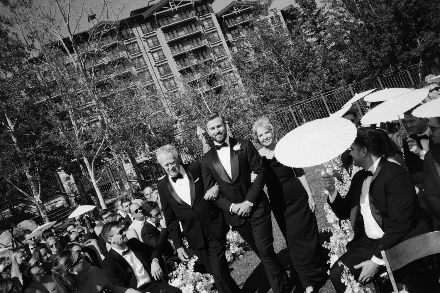 Black and white photo of wedding party walking in a line holding umbrellas during an outdoor celebration.