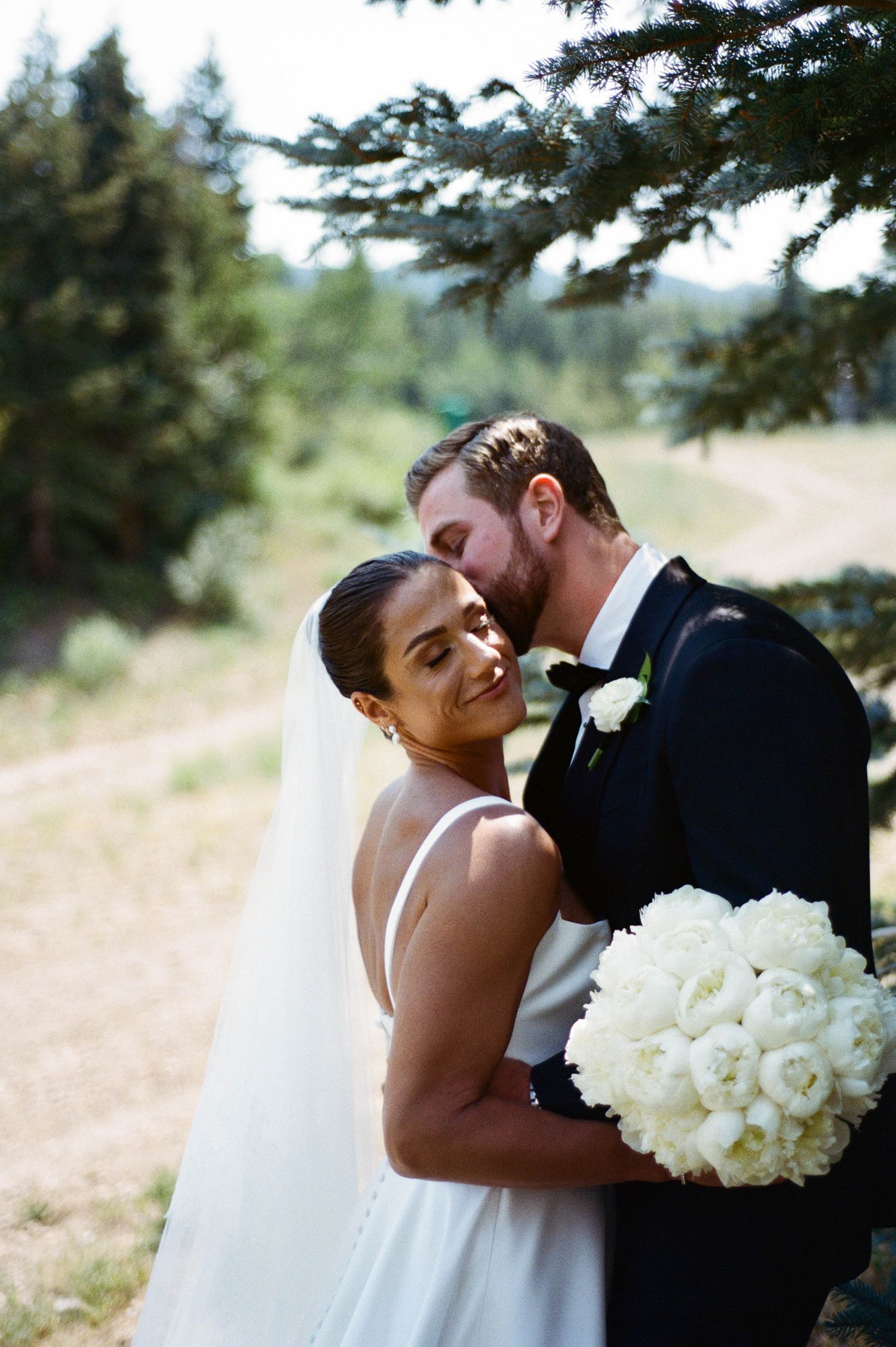 Romantic wedding couple embraces outdoors with white bouquet and evergreen trees in background.