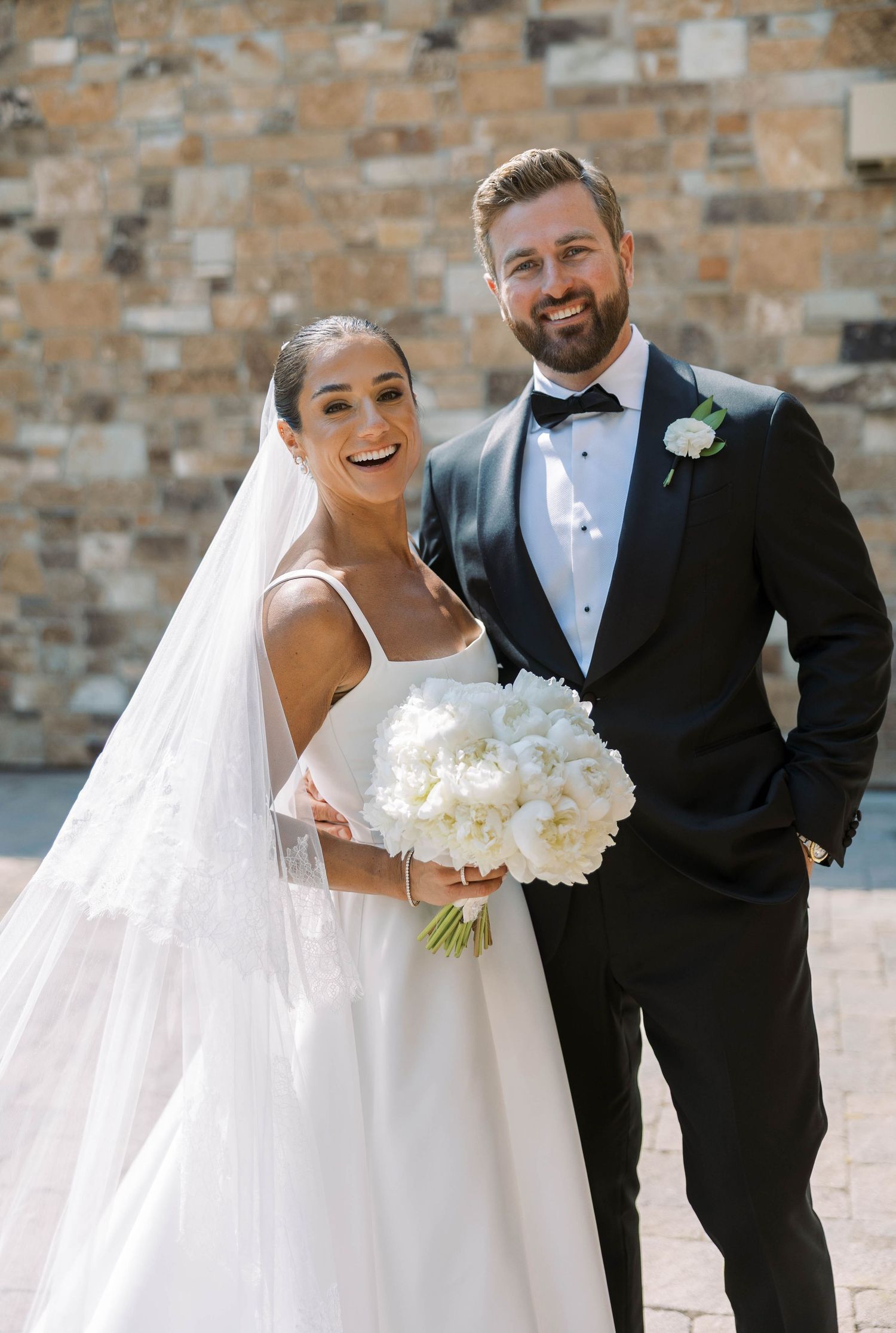 Wedding couple poses against rustic brick wall while sharing laughs and holding white bouquet.