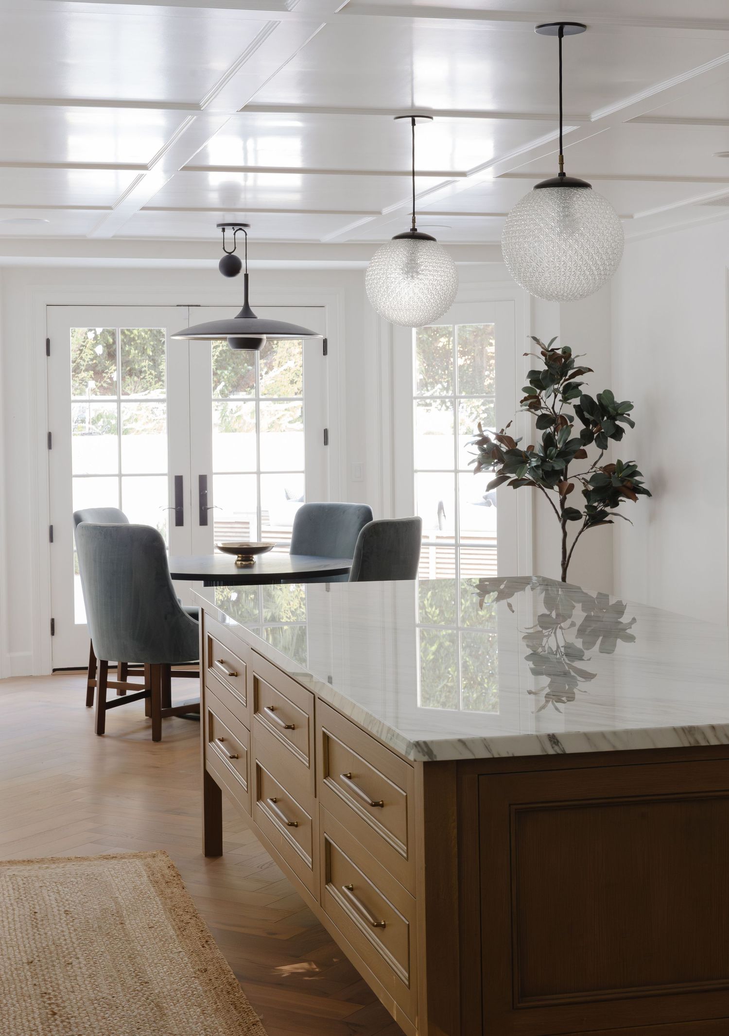 Modern kitchen island with marble countertop, warm wood cabinetry, globe pendant lights, and French doors leading to a bright dining area.