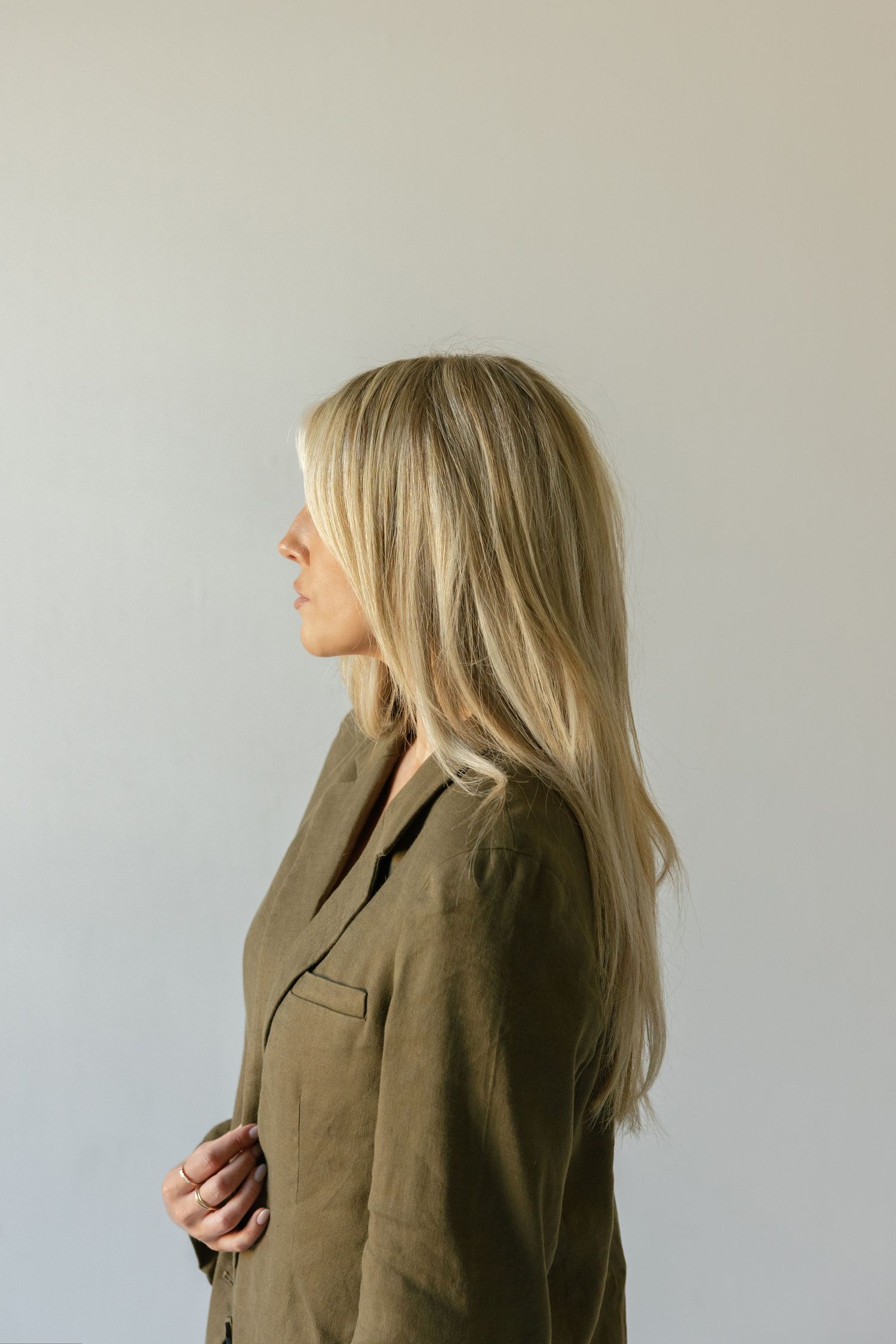 Woman with long blonde hair in profile view wearing an olive green linen blazer against a light gray background.