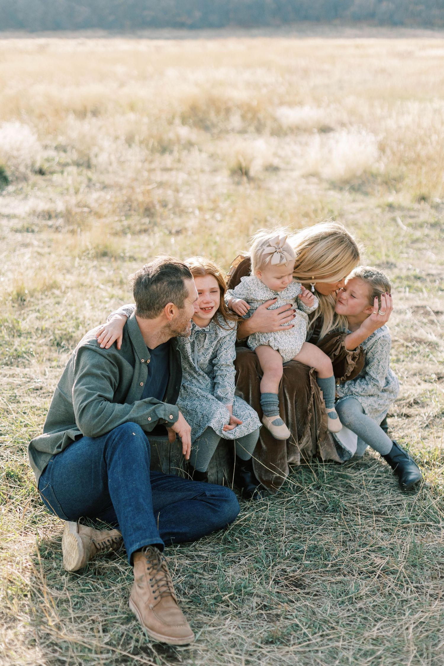 A family shares an intimate moment while sitting in tall grass with mountains behind them.