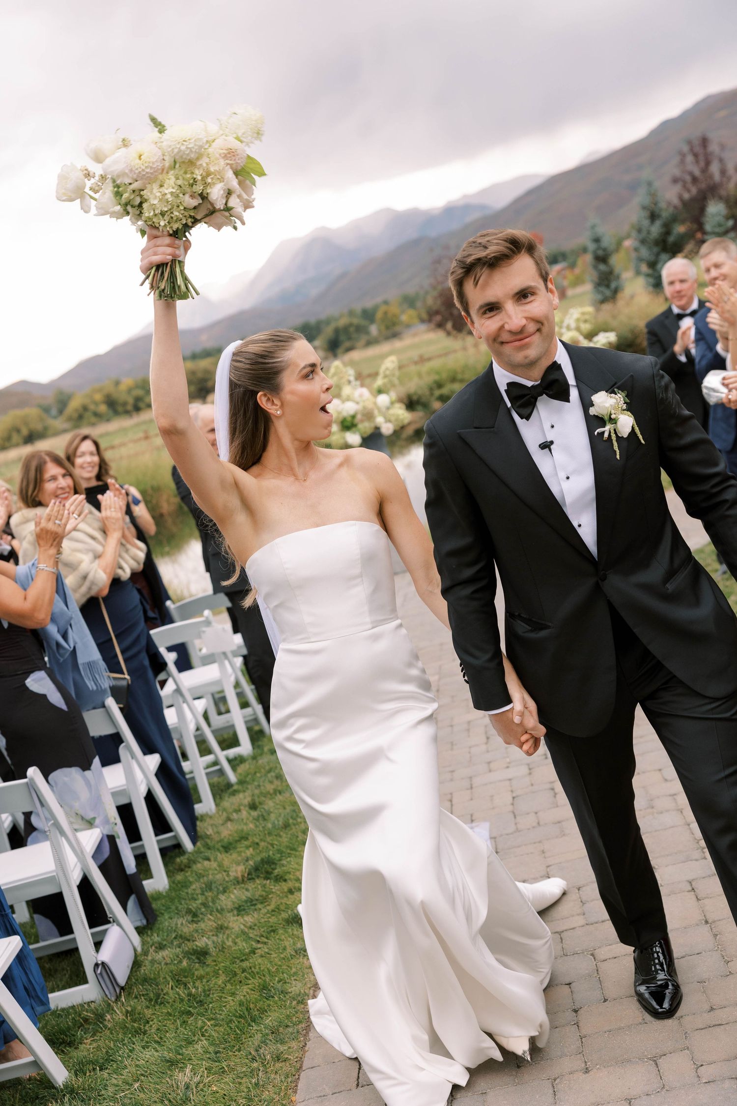 Wedding couple celebrates while walking down stone pathway with mountain backdrop and white chairs.