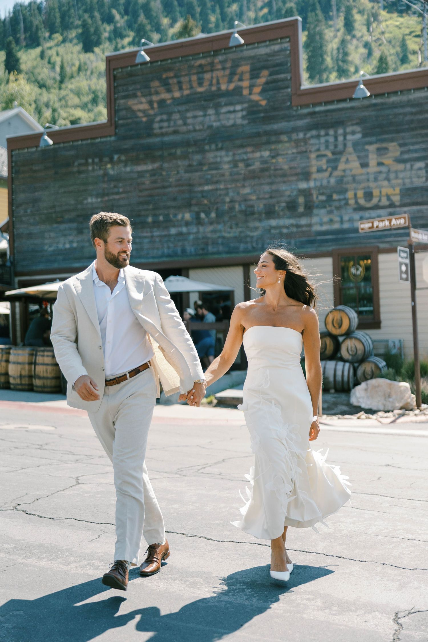 A couple in white wedding attire walks hand in hand in front of a rustic storefront on a sunny day.