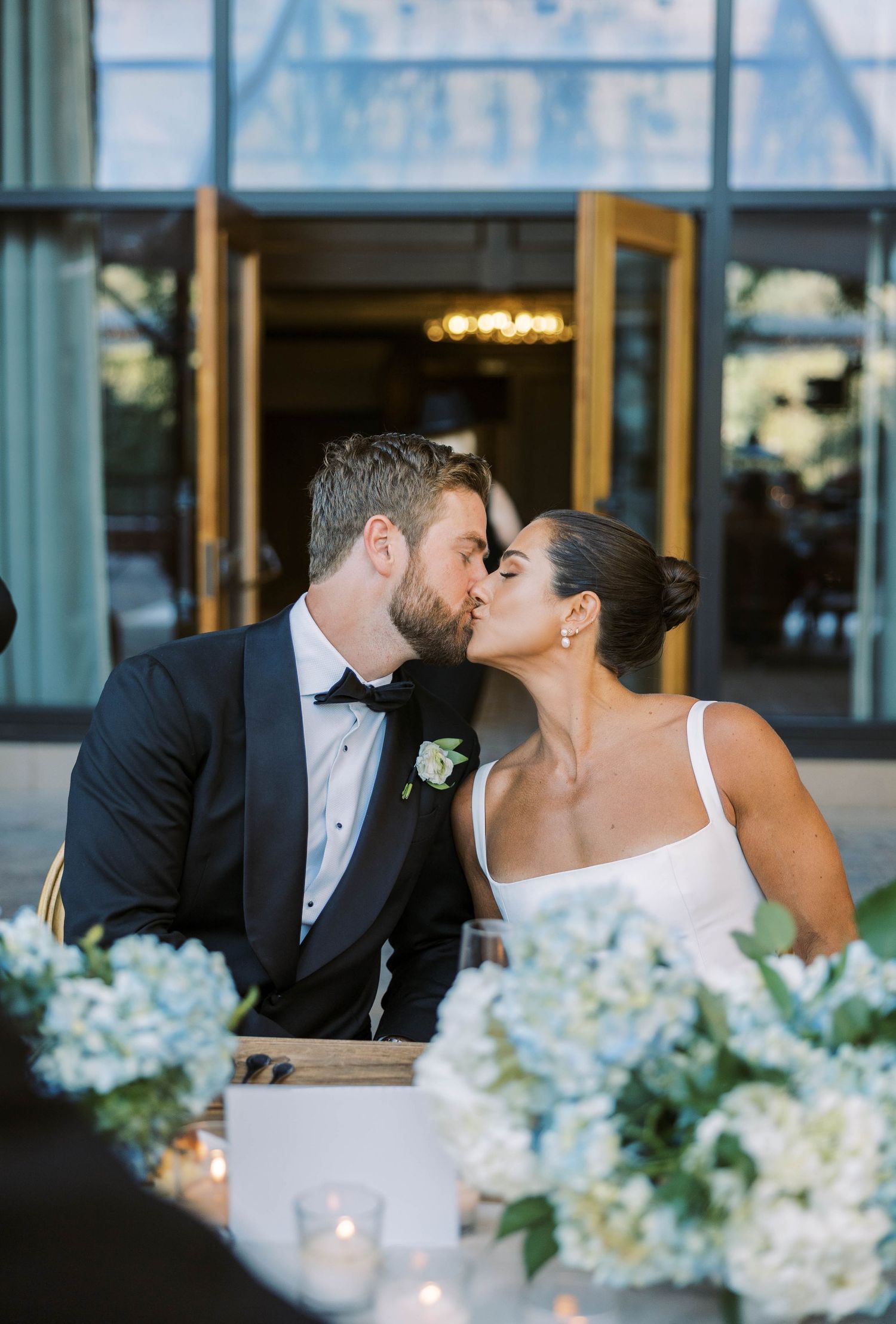 A romantic wedding couple shares a kiss at an elegant reception with blue hydrangea centerpieces and candlelight.