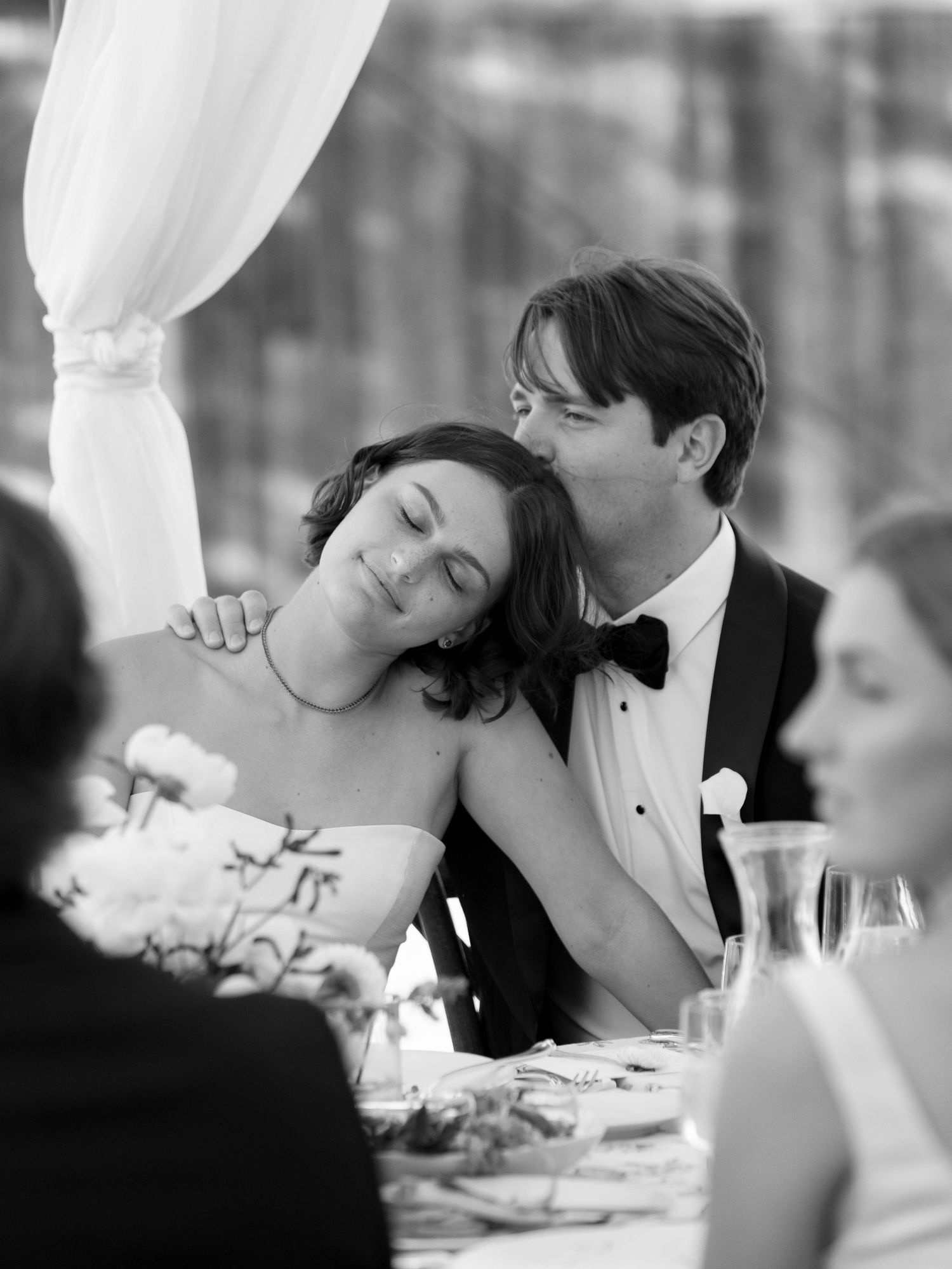 A romantic black and white wedding reception scene showing a tender moment between a couple at their dining table.
