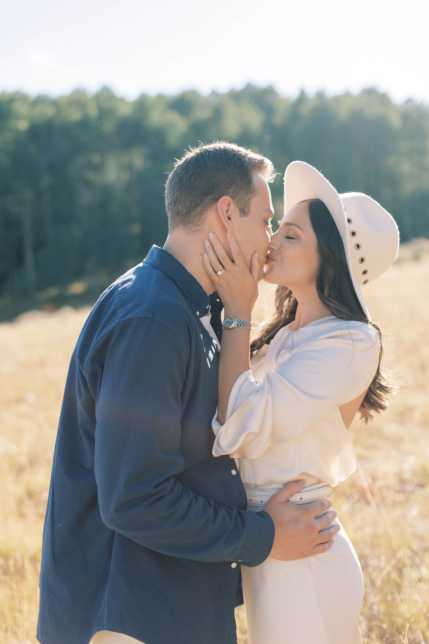 Romantic kiss shared in sunlit mountain meadow with white hat detail.