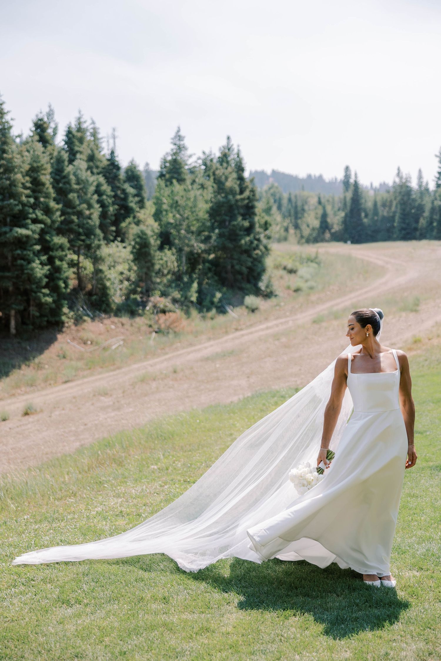 A bride's dress flows gracefully across a mountain meadow with evergreen trees in the background.