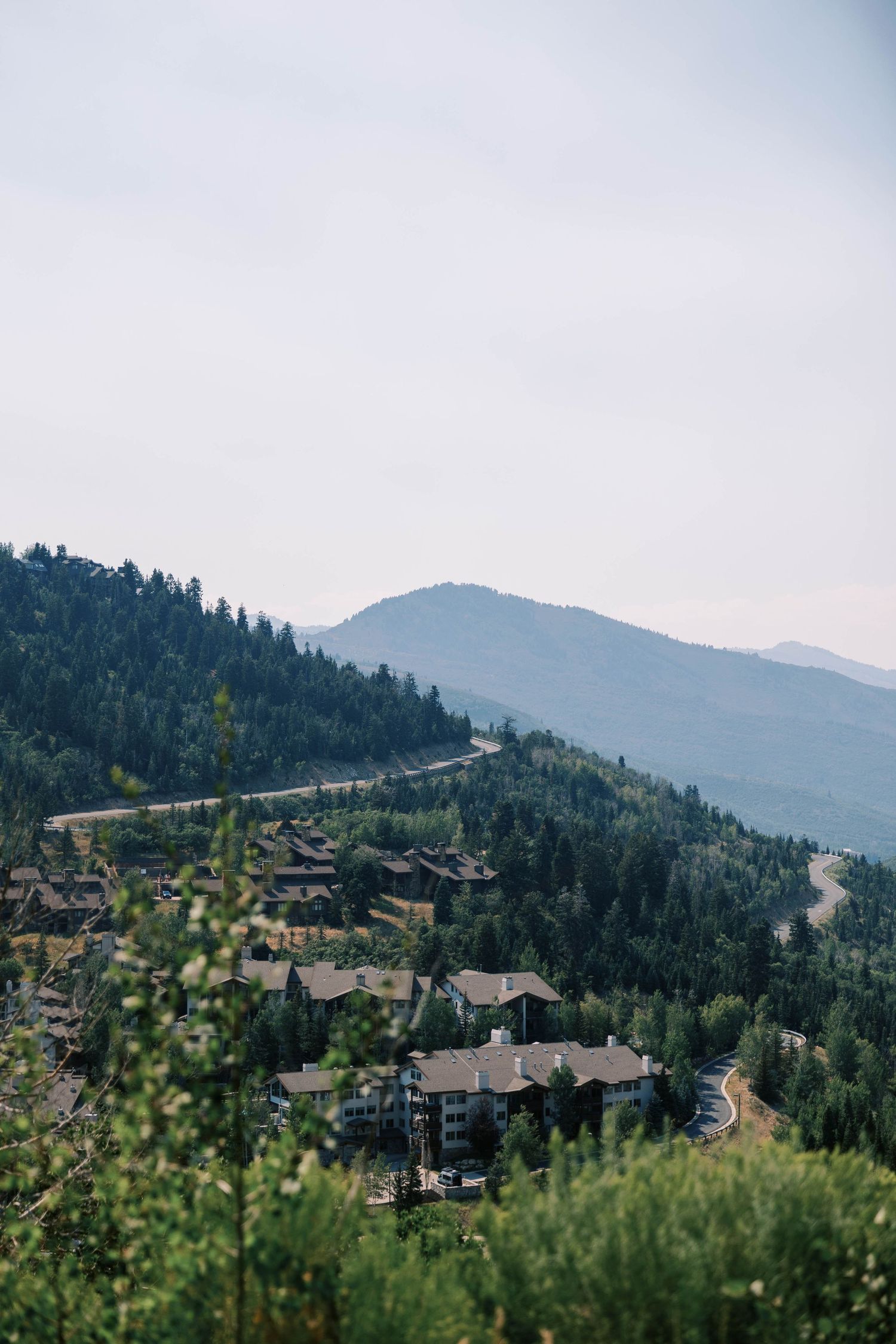 A winding mountain road curves through a forested hillside with lodges nestled among pine trees in a scenic landscape.