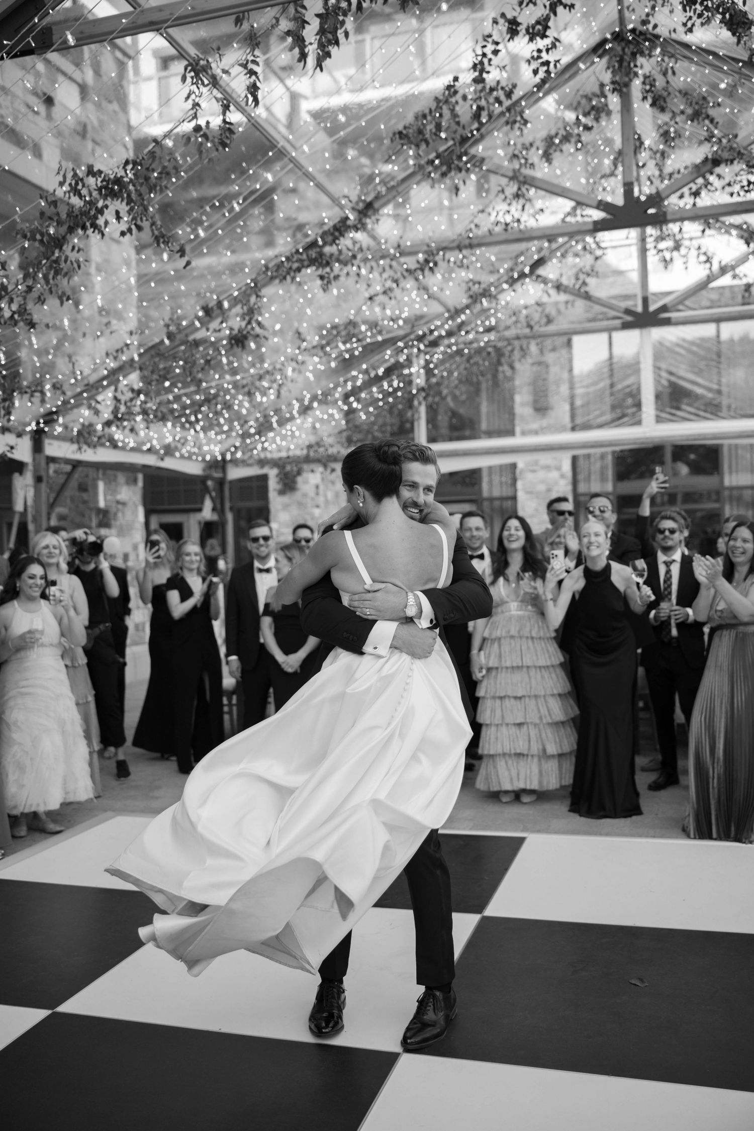 A couple dances on a black and white checkered dance floor under twinkling lights at an elegant wedding reception.