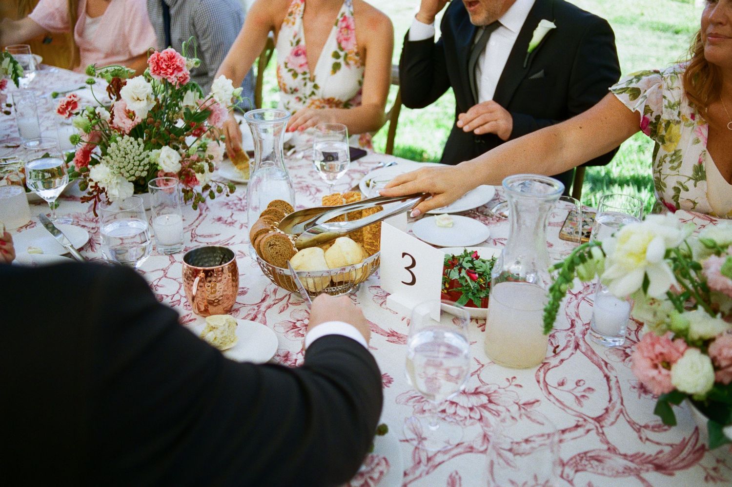 Hands serve food at an elegant wedding reception table decorated with pink floral arrangements and crystal glasses.