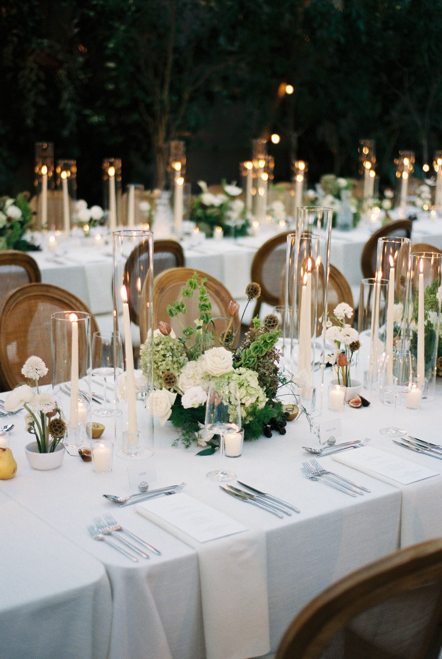 Elegant wedding reception table settings with white linens, tall candles, and delicate greenery centerpieces.