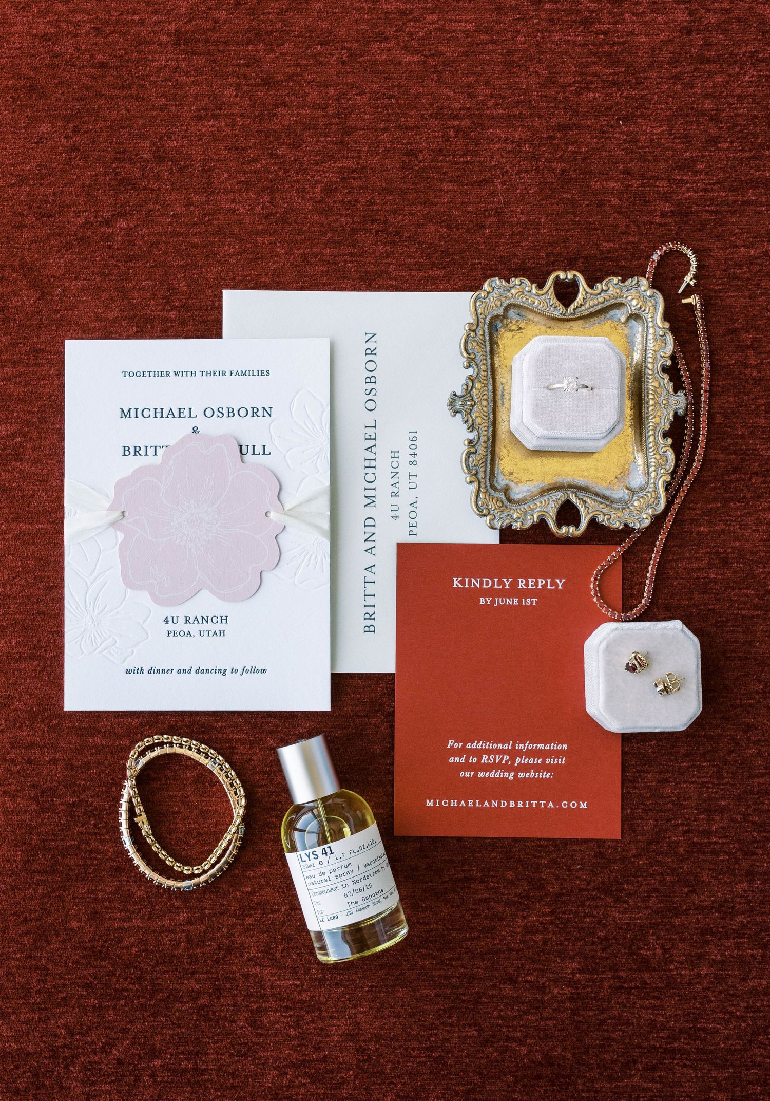 Elegant wedding invitation suite with gold accents, jewelry, and perfume bottle arranged on deep red velvet background.