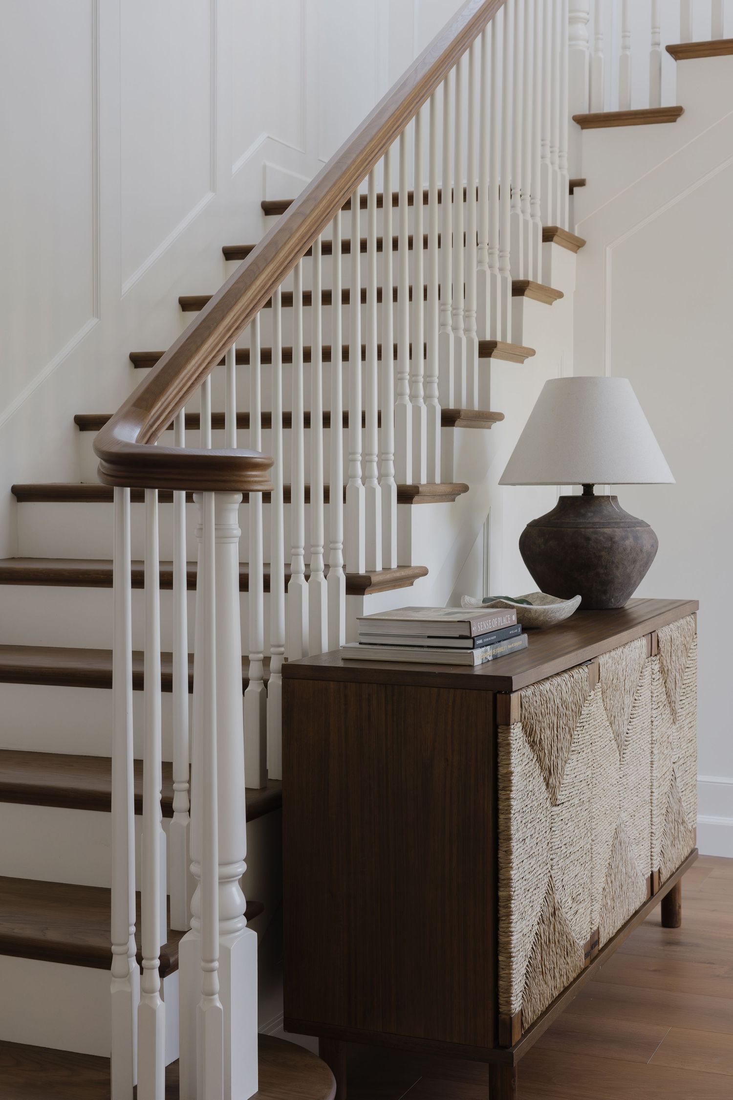 Elegant entryway with white spindle staircase, dark wood steps, and a textured sideboard topped with a rustic ceramic lamp.