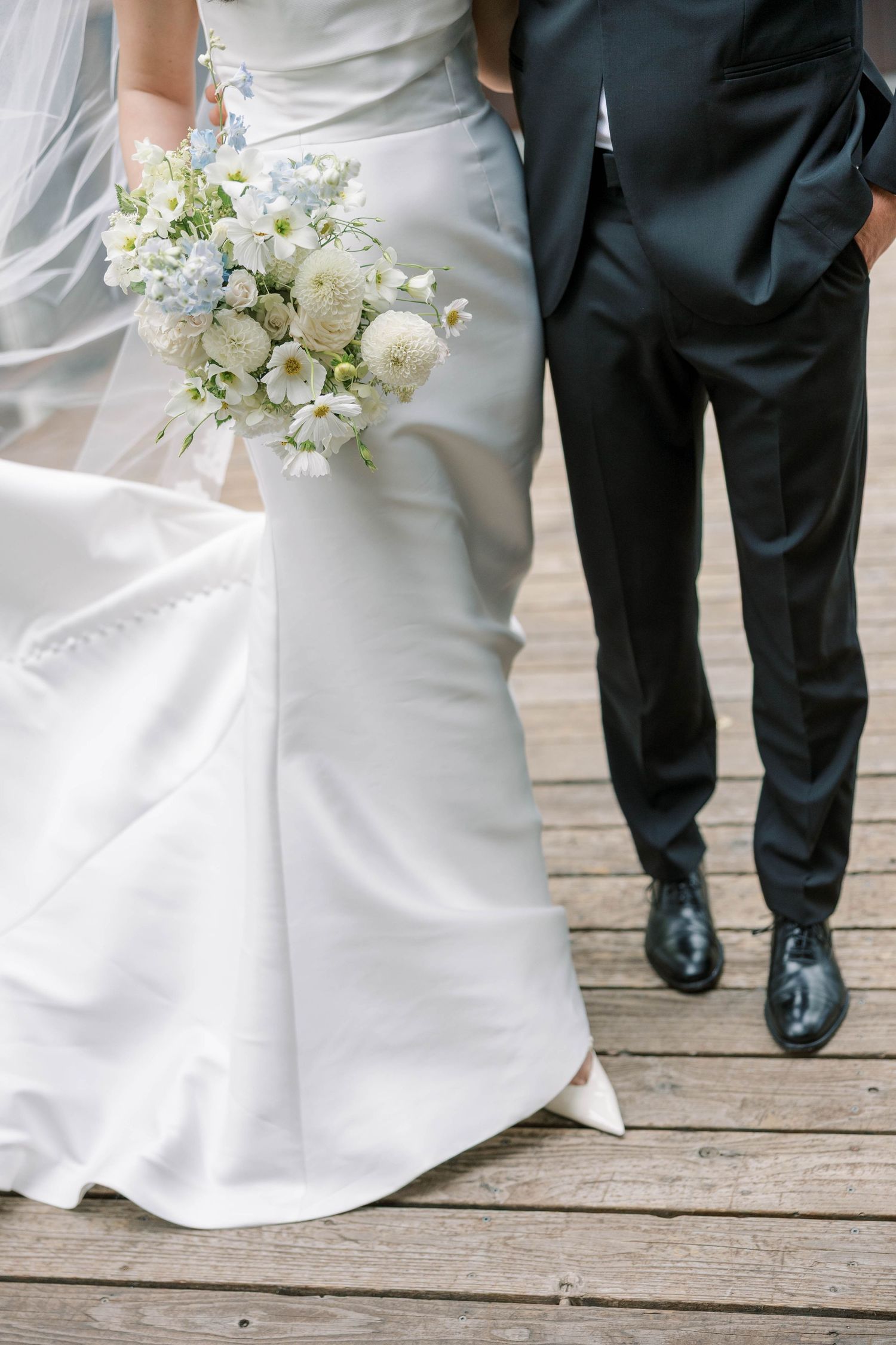 Close-up details of a white wedding dress and black tuxedo shoes on a wooden deck.