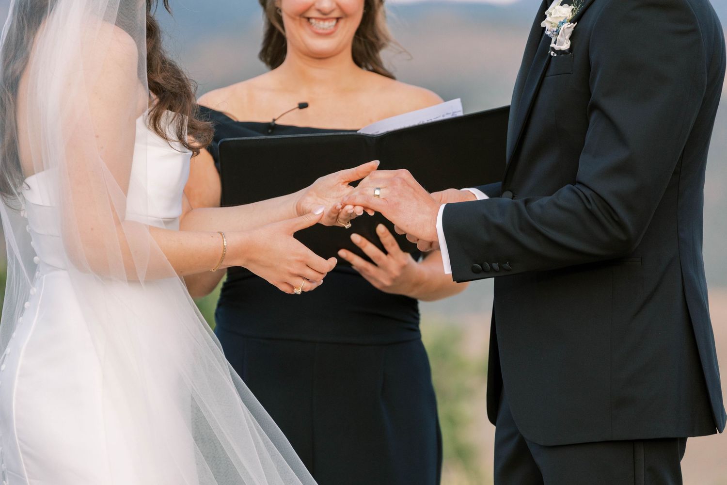 A couple holds hands during an outdoor wedding ceremony with mountains visible in the background.