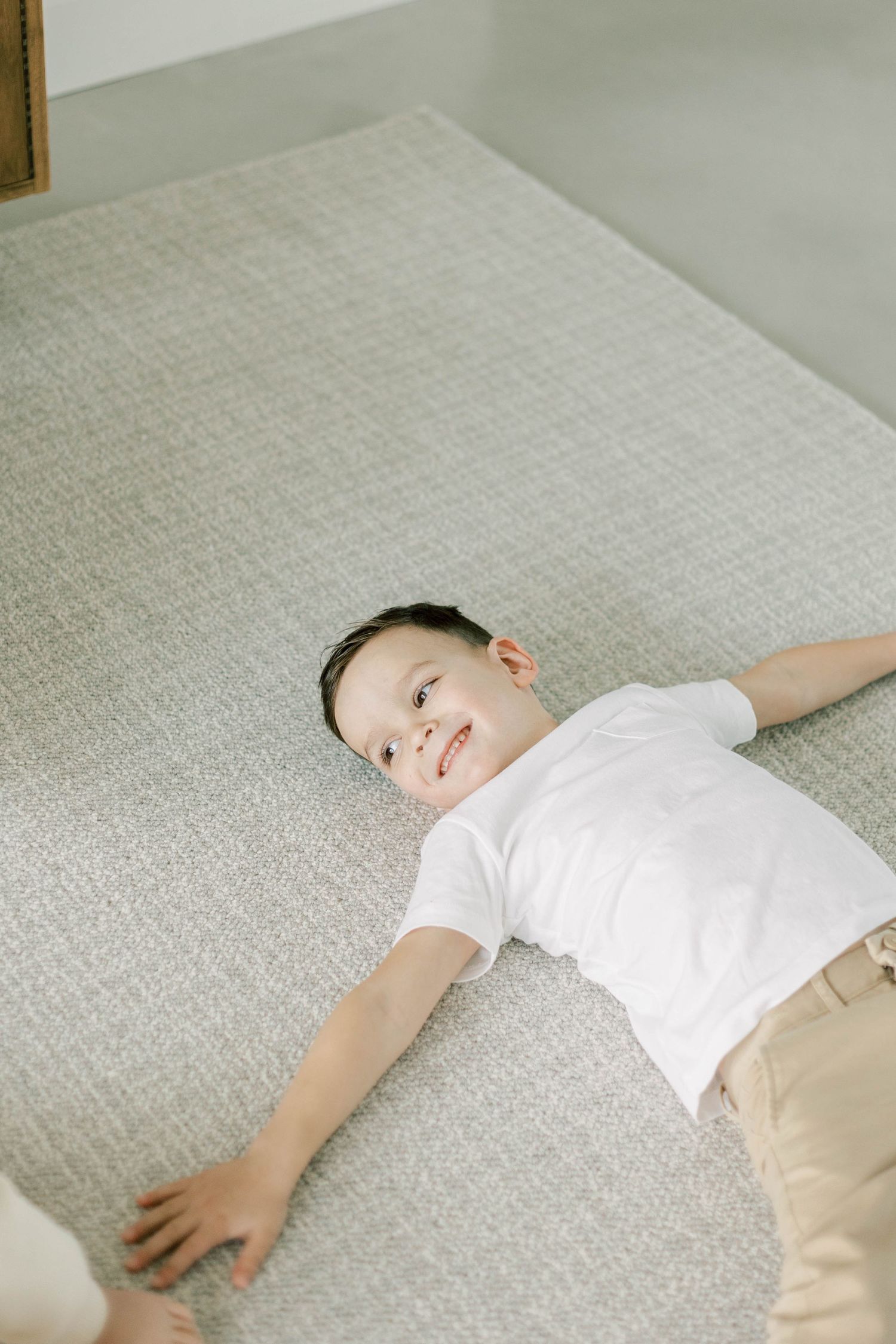 Young child in white t-shirt and khaki pants lying on gray upholstered surface.