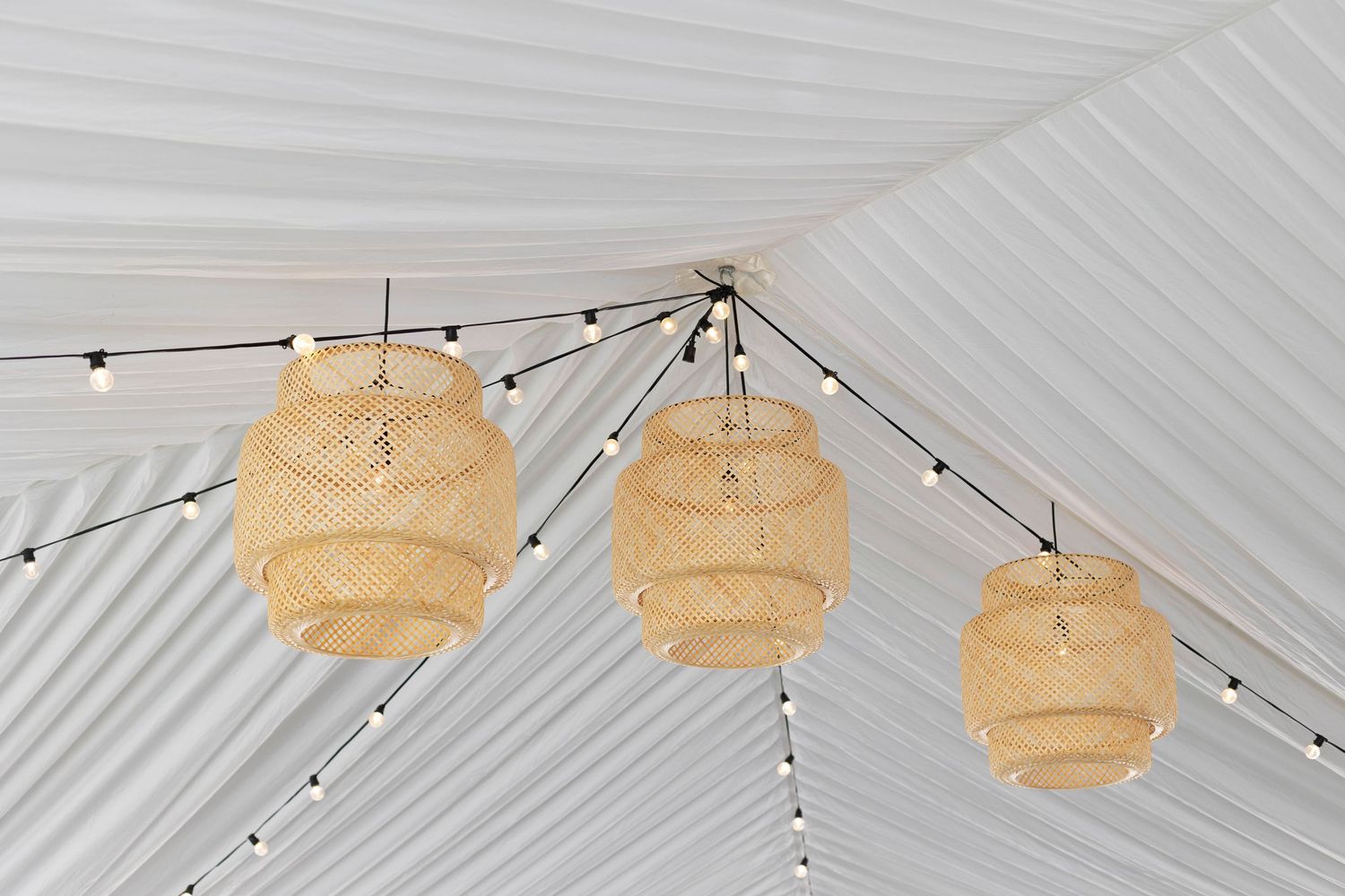 Woven bamboo pendant lights hanging from white tent ceiling with string lights.