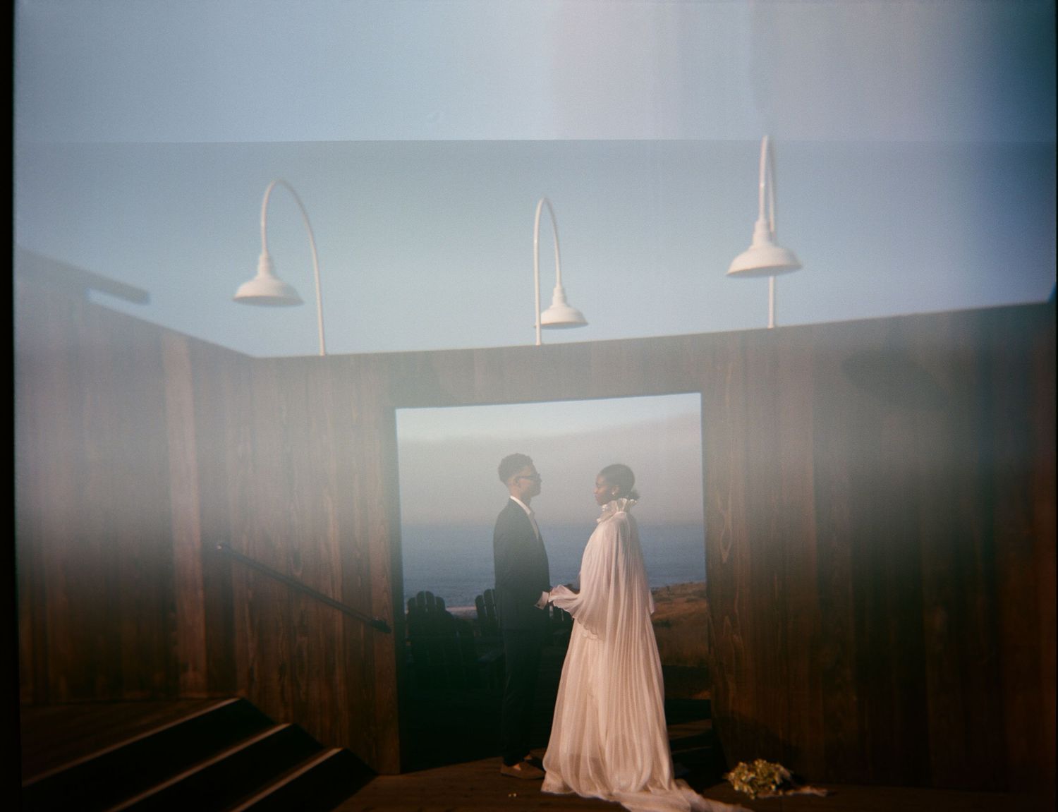 Two silhouettes stand at a window with pendant lights hanging above in a moody, atmospheric interior setting.