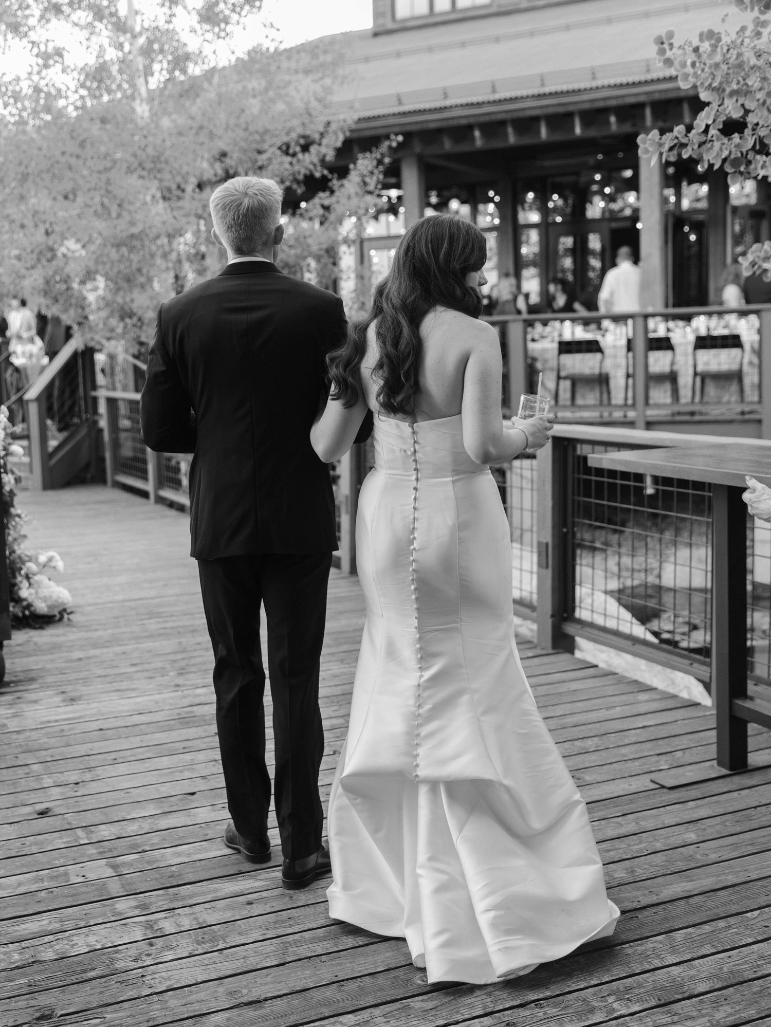 A couple in formal wedding attire walks along a wooden deck towards a rustic venue.