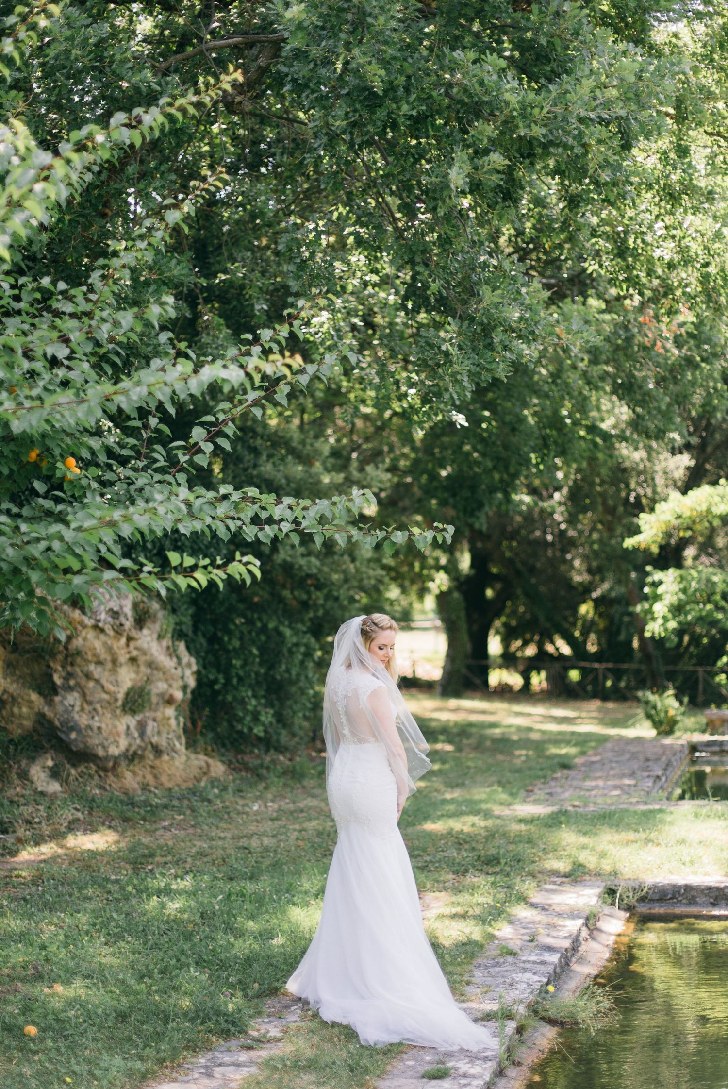 Full-length bridal gown silhouette beside a garden pond with trees in background.