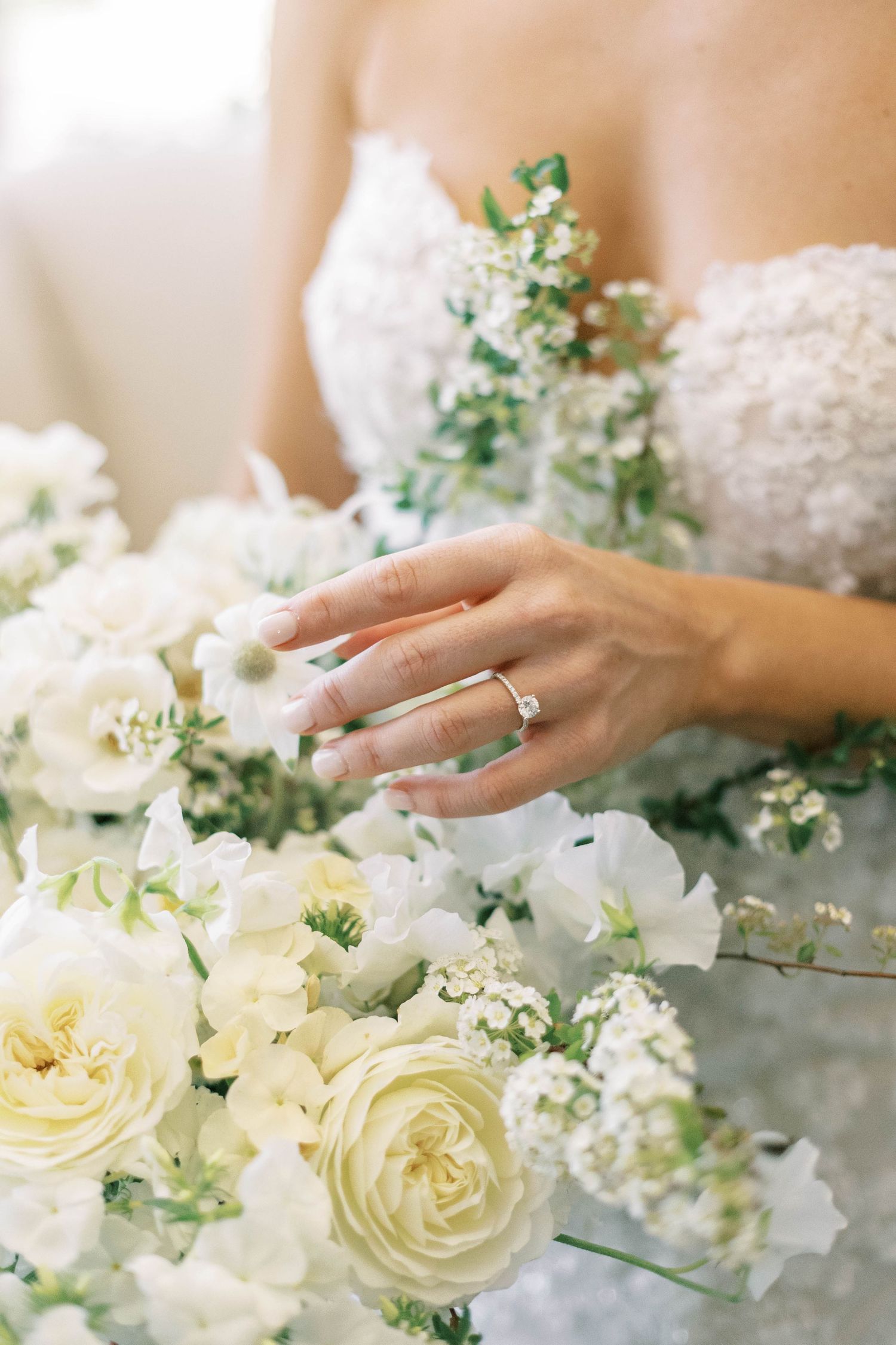 Close-up of bride's hand with wedding ring among white roses and delicate floral arrangements.