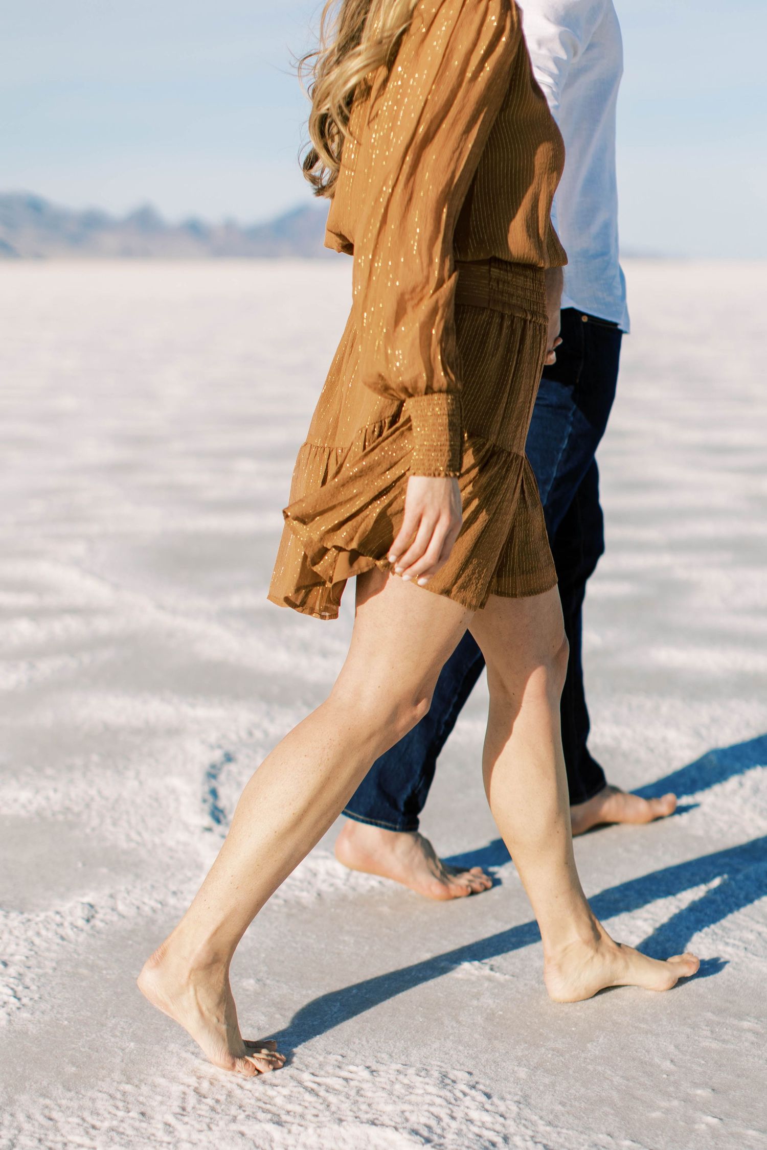 Close-up of legs walking on white salt flats wearing camel dress and dark jeans.
