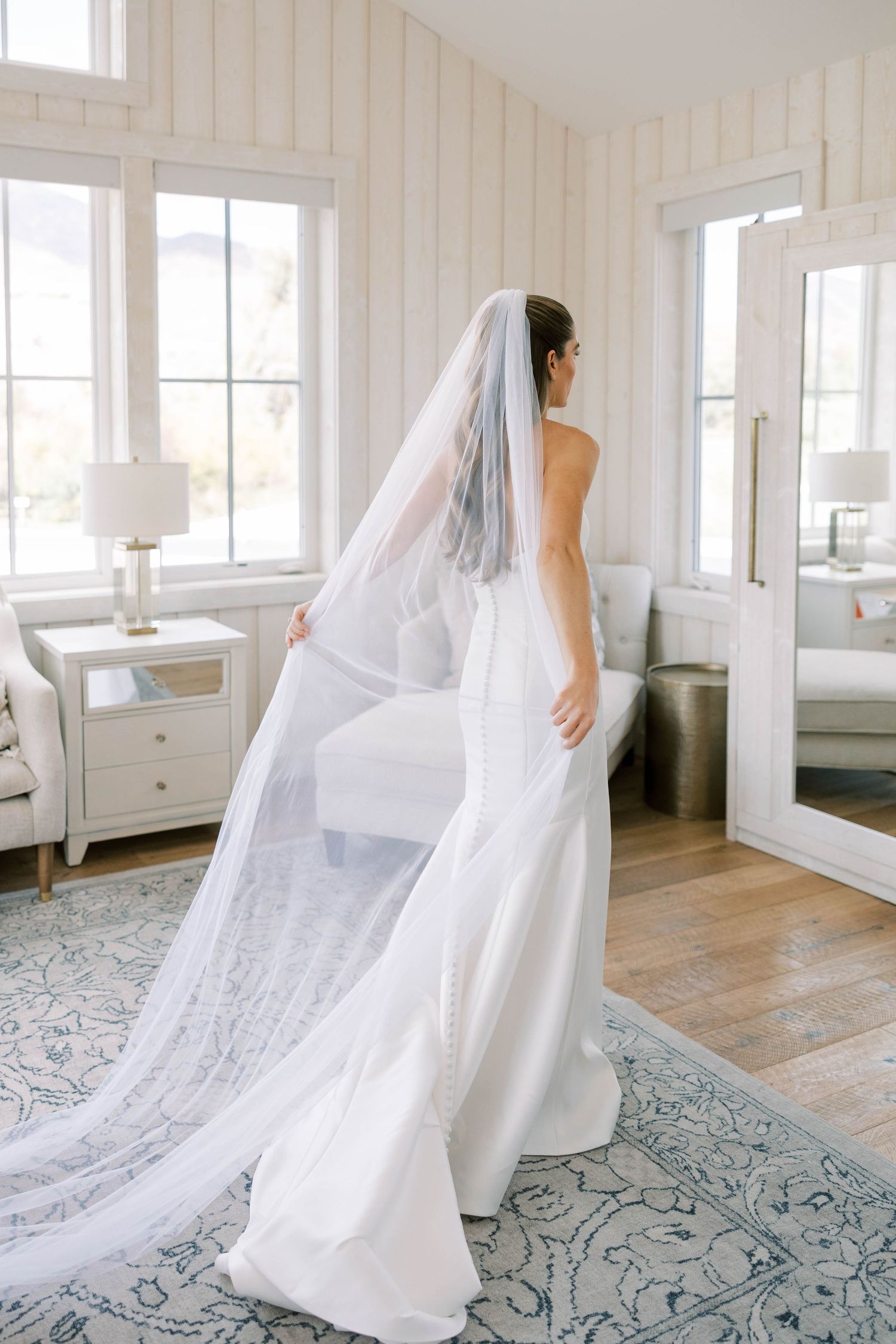 A flowing white wedding veil trails behind a bride in an elegant white dress standing in a bright sunlit room.