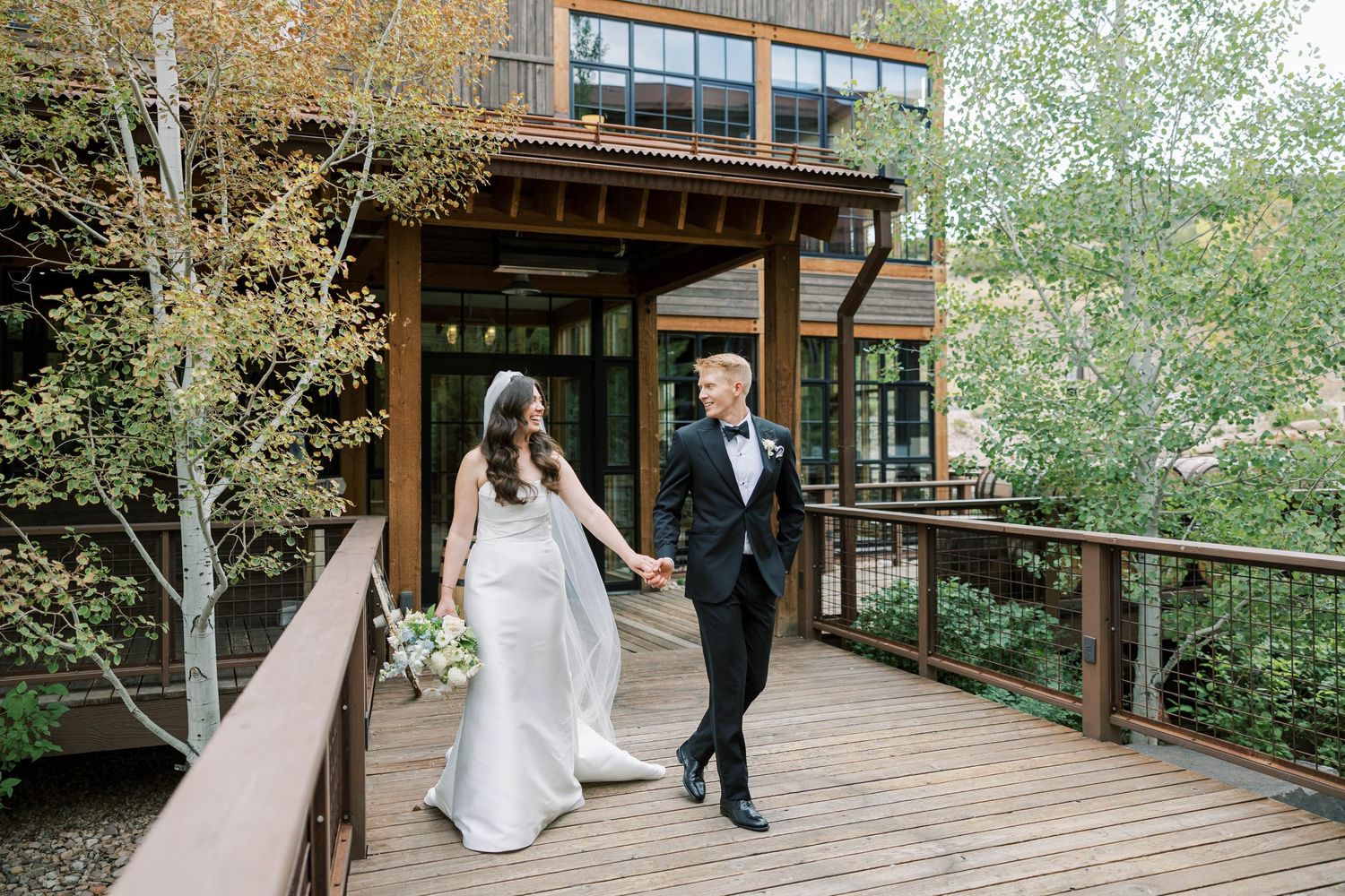 Couple in formal attire walking together along a wooden deck surrounded by lush greenery on their wedding day.