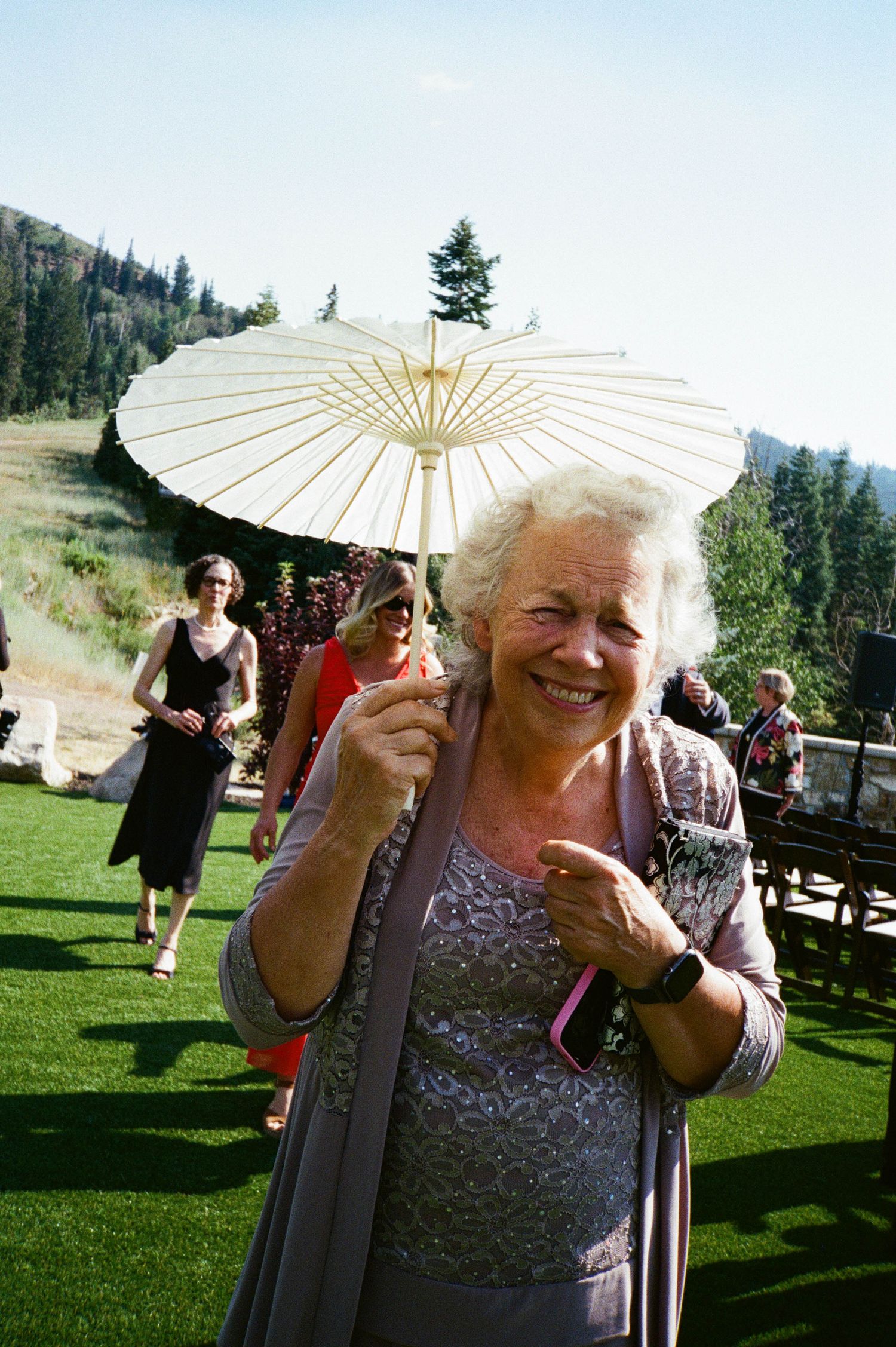 A parasol-carrying guest smiles during an outdoor summer celebration on a grassy lawn with mountains in the background.